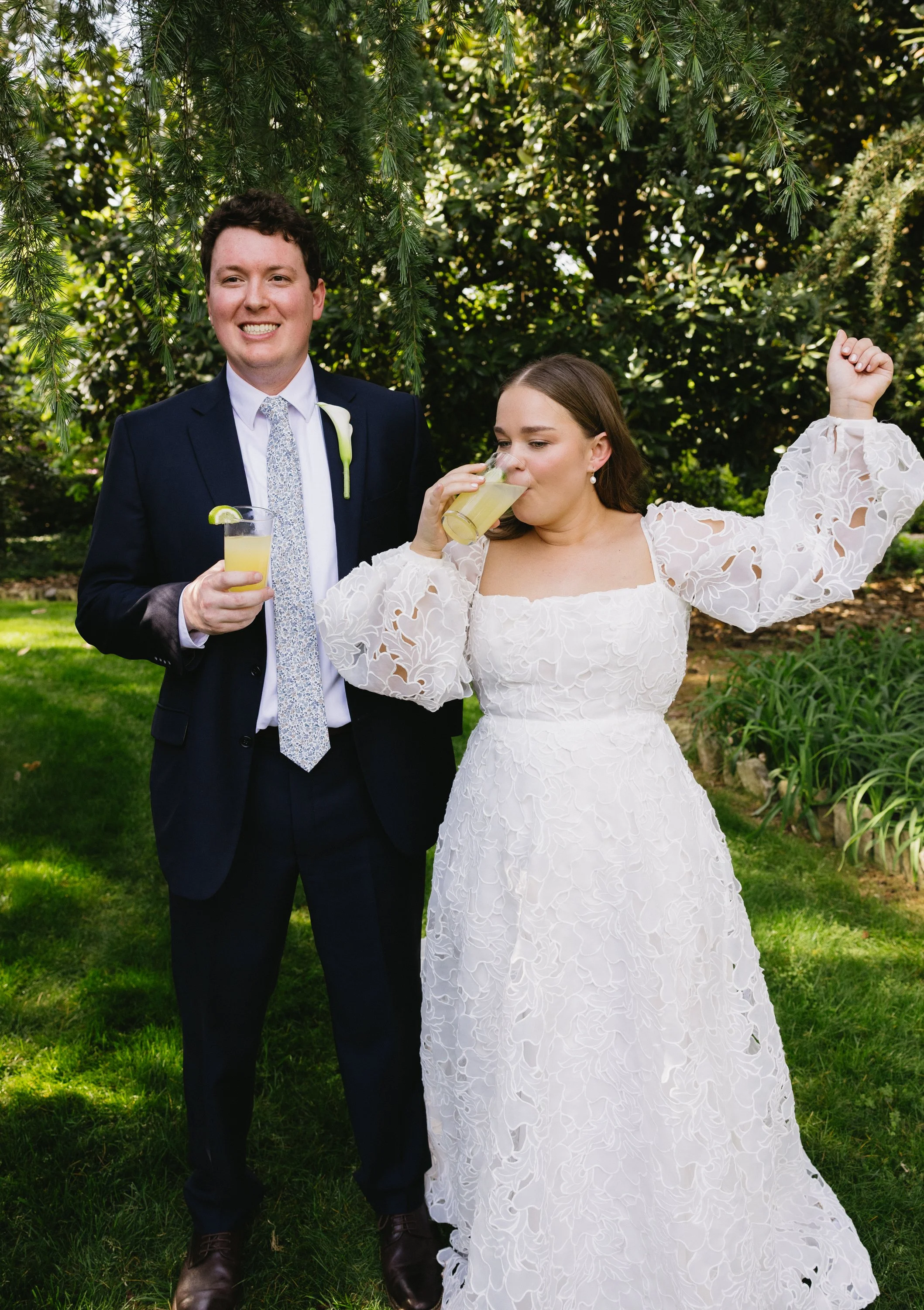 A newlywed couple standing outdoors under a tree, enjoying drinks. The groom is in a dark suit with a patterned tie, holding a drink with a lime wedge. The bride is in a white lace wedding dress, sipping her drink, with one arm raised in celebration.