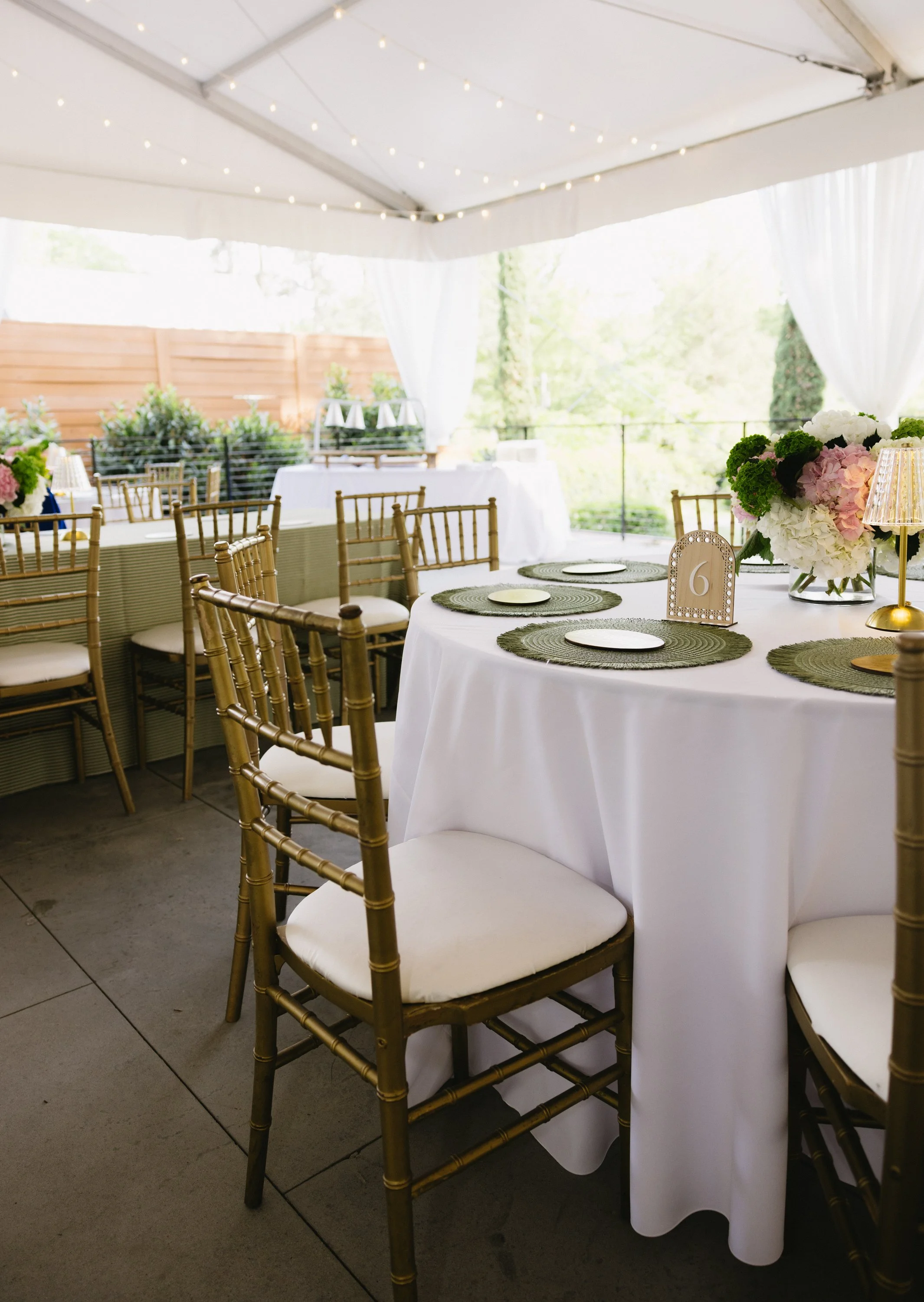 Elegant outdoor event tent with white drapes and string lights, decorated with round tables covered in white tablecloths, gold chairs, floral centerpieces, and table numbers.