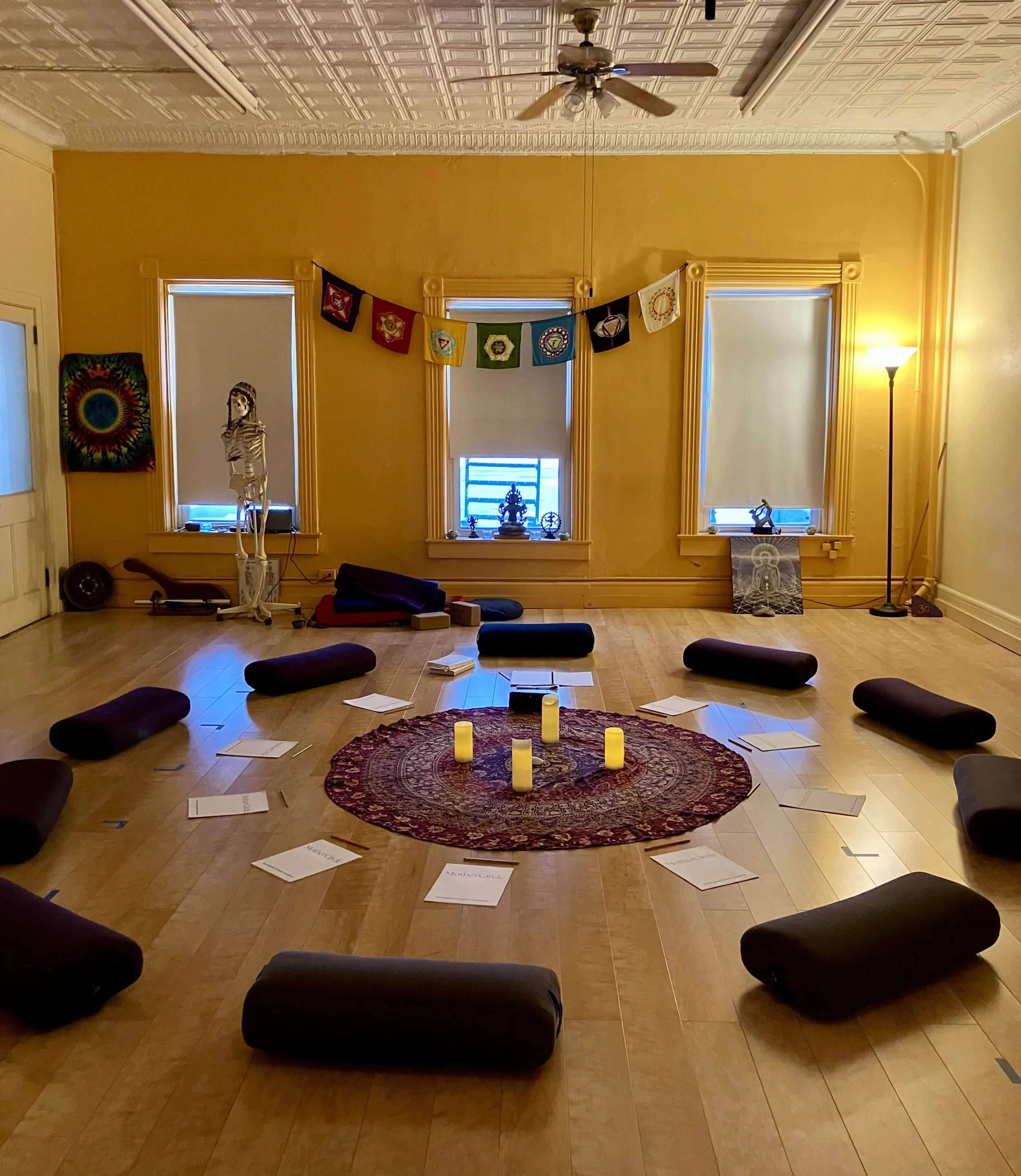 A yoga or meditation space with a circular arrangement of black cushions and candles on a decorative rug, surrounded by spiritual and artistic decorations against yellow walls with large windows.