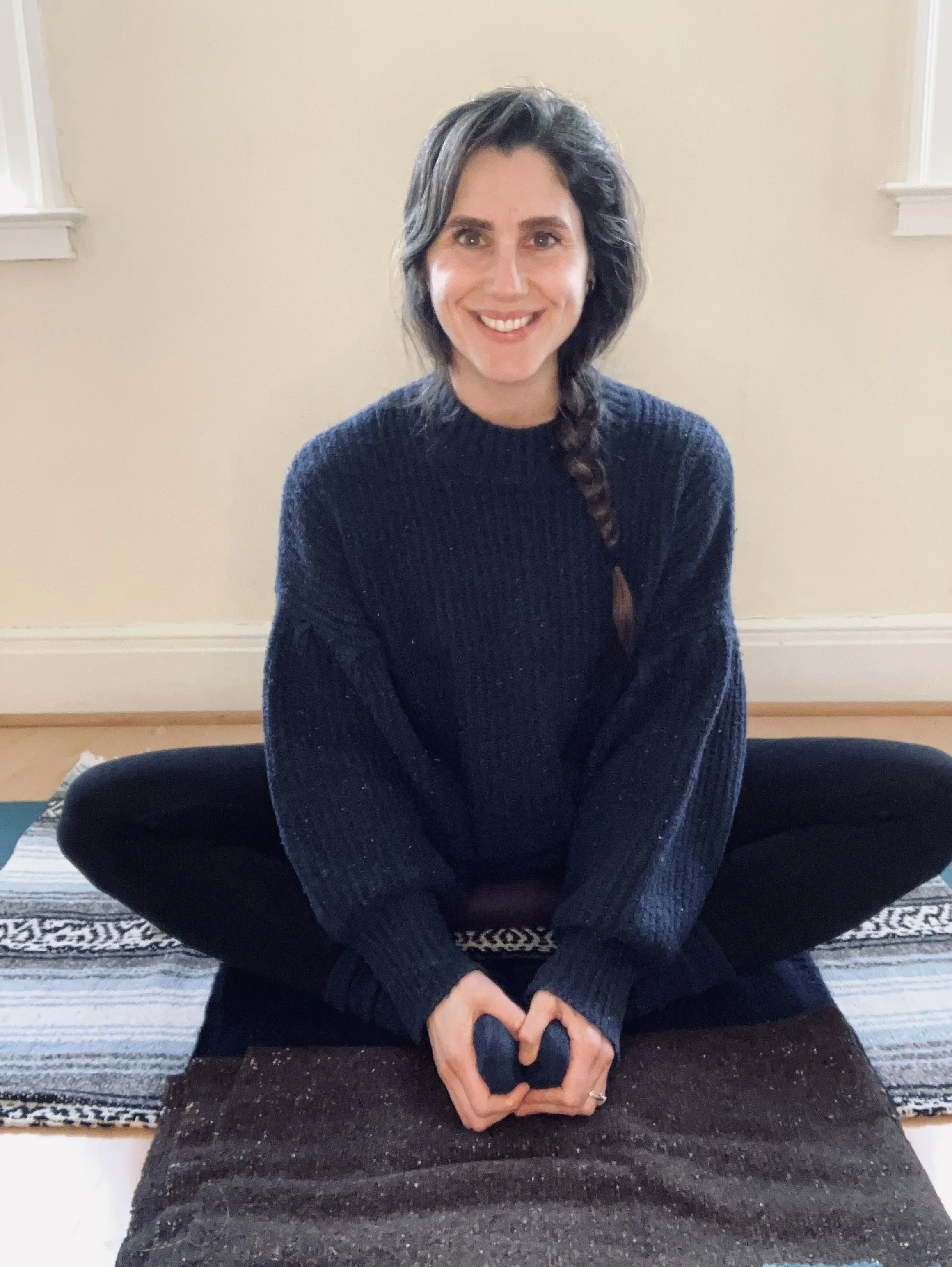 A woman with shoulder-length dark hair and a braid, sitting cross-legged on a yoga mat, smiling at the camera in a room with beige walls and two small windows.