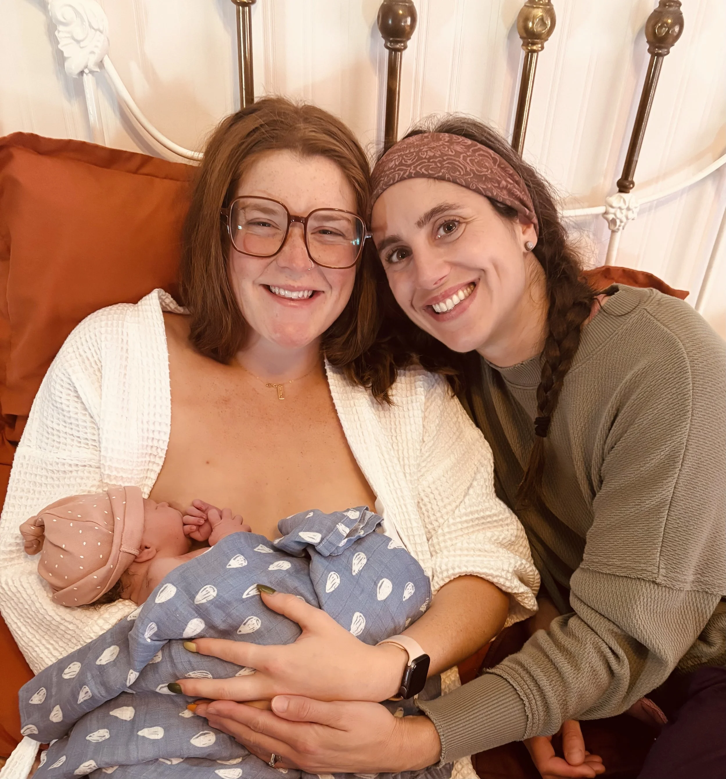 Two women, one holding a newborn baby, all smiling, in a cozy indoor setting with a metal headboard and rust-colored pillows in the background.