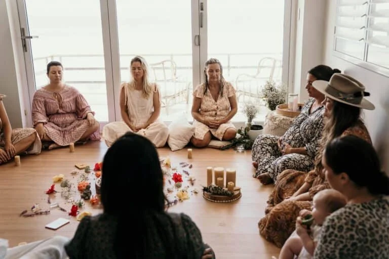 Group of women sitting cross-legged in a circle inside a bright room, with candles and flowers in the center.
