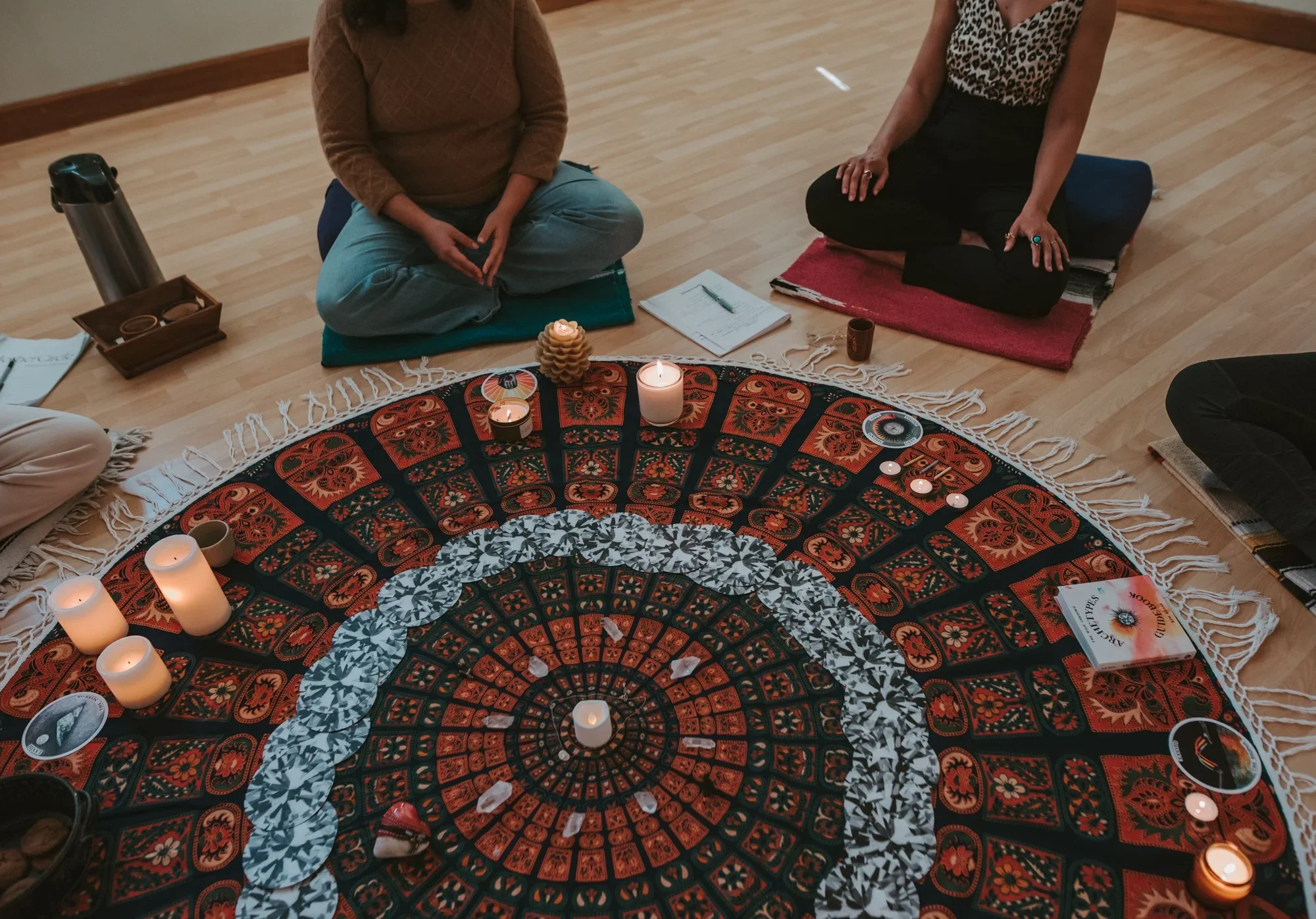Two women sitting cross-legged on cushions around a decorated circular cloth with candles and crystals, participating in a wellness or spiritual gathering.