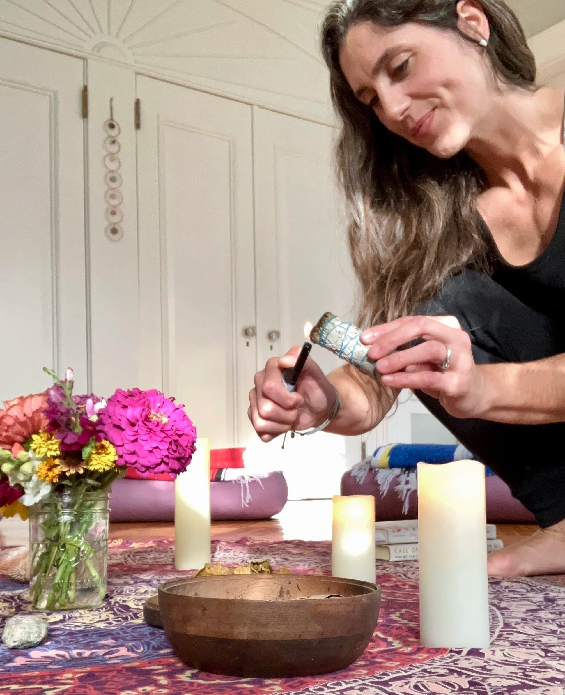 Woman lighting a candle on a table with flowers, candles, and books.