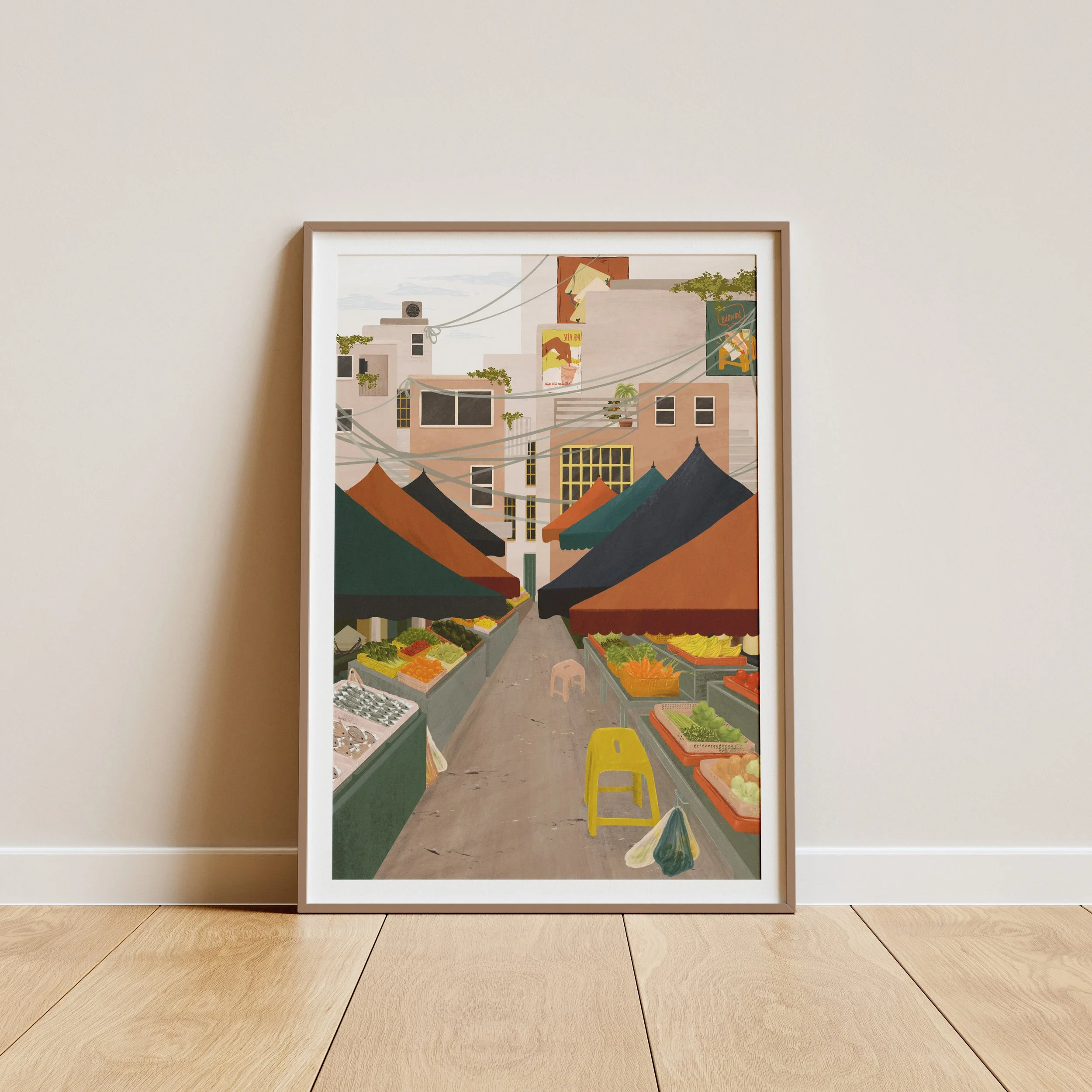 Framed colorful artwork of a street market scene with produce stalls, tents, and buildings in the background, leaning against a wall on a wooden floor.