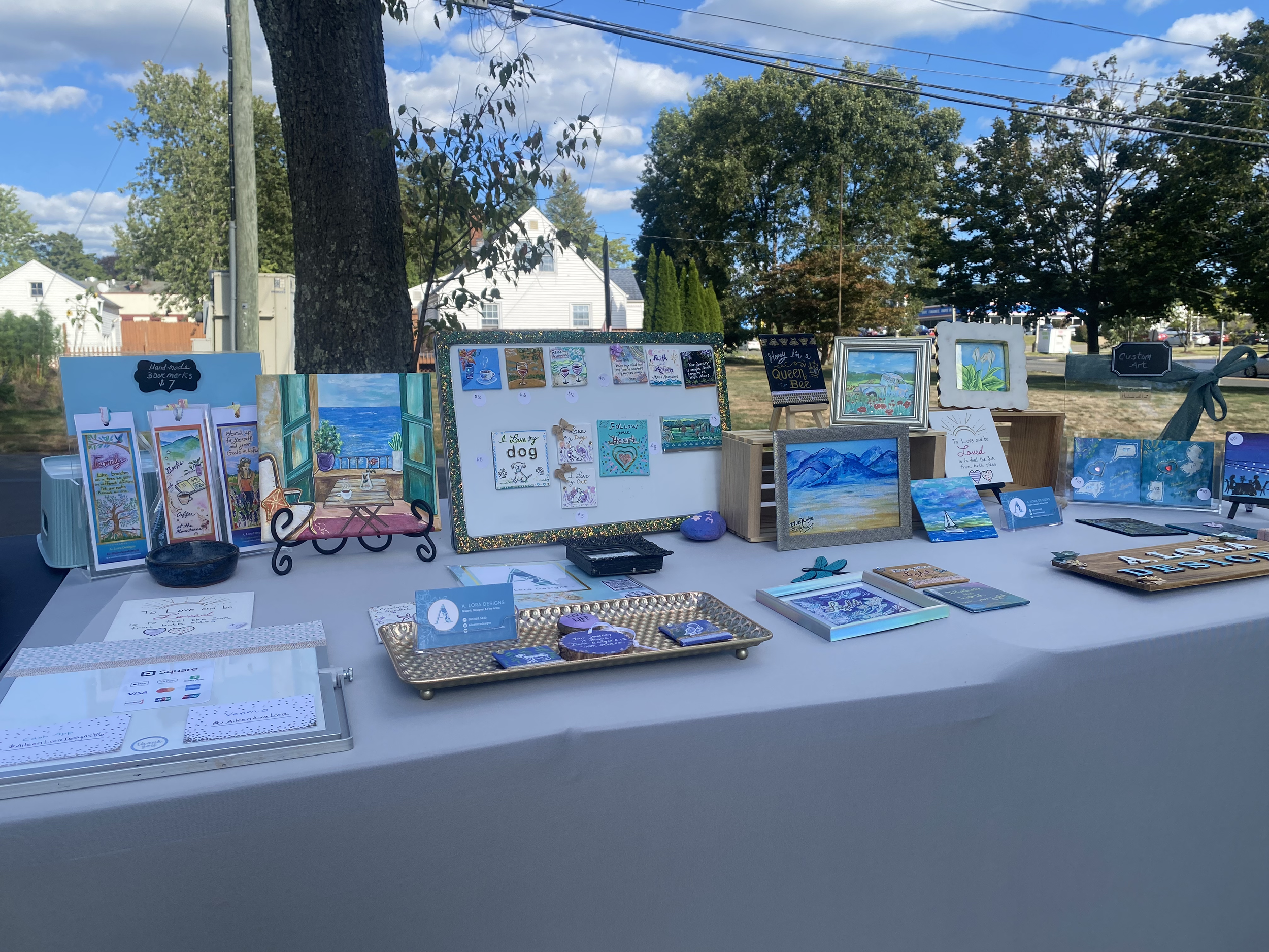Outdoor art and craft display table featuring paintings, cards, and handmade items, set against a backdrop of trees, houses, and a blue sky.