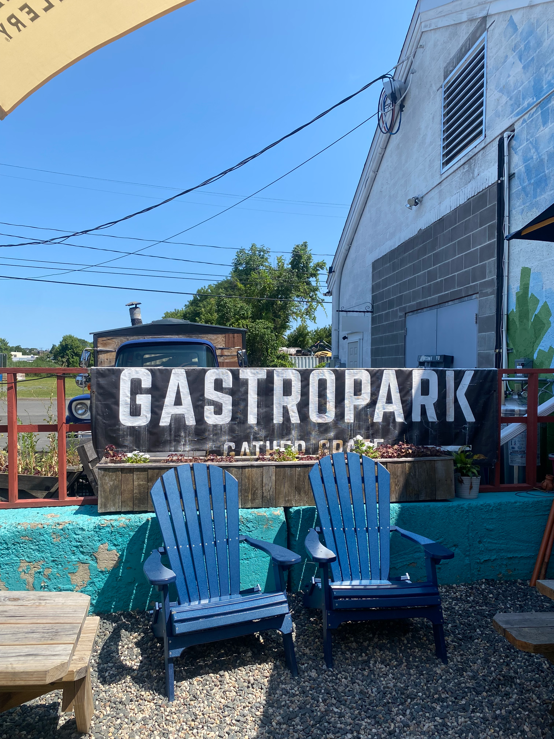 Outdoor seating area with two blue Adirondack chairs in front of a banner that reads 'GASTROPARK' on a building with a white and gray exterior, under a clear blue sky.
