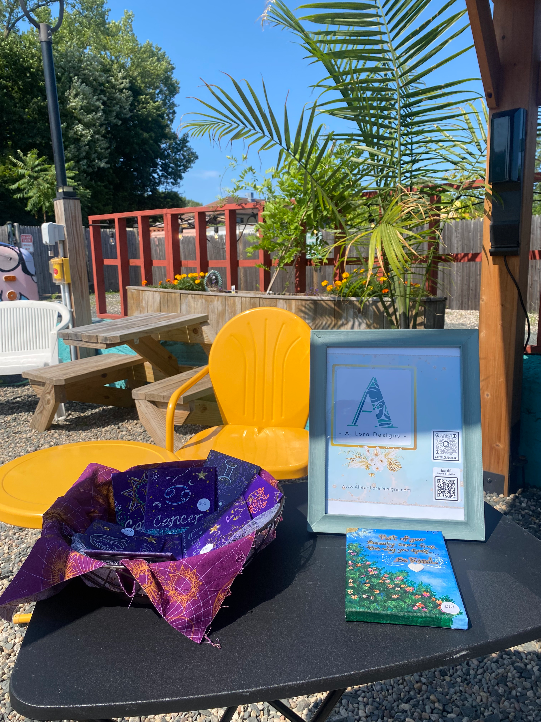 Table with purple bandanas featuring astrology signs, a framed sign with a logo and contact information, and a flyer that says 'Part of your beauty comes from the way you speak. Be Kind.' outside area with yellow chairs, wooden picnic table, and plan