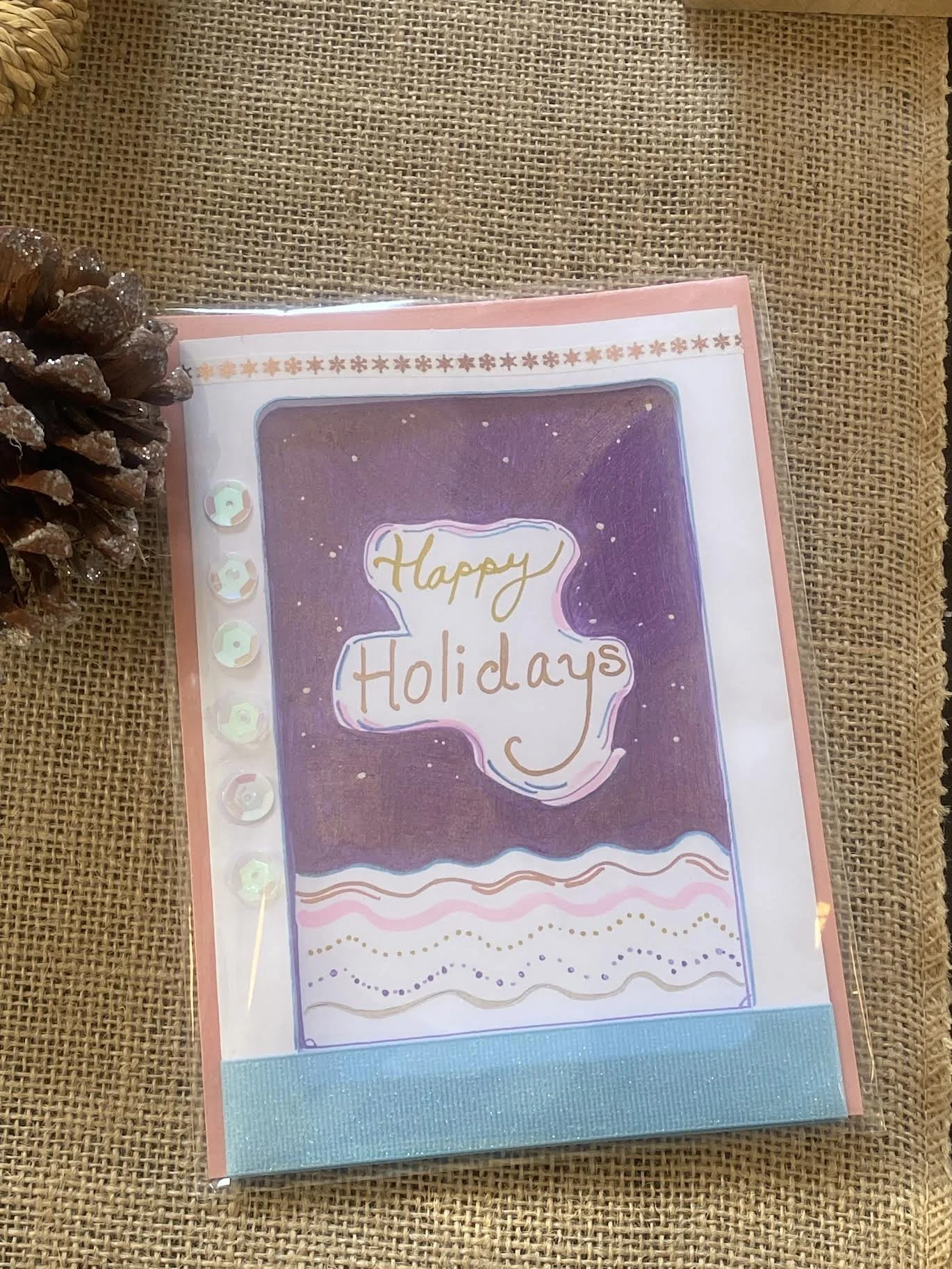 A greeting card with a purple background, decorated with gold, pink, and white doodles and the words 'Happy Holidays' in handwritten style.