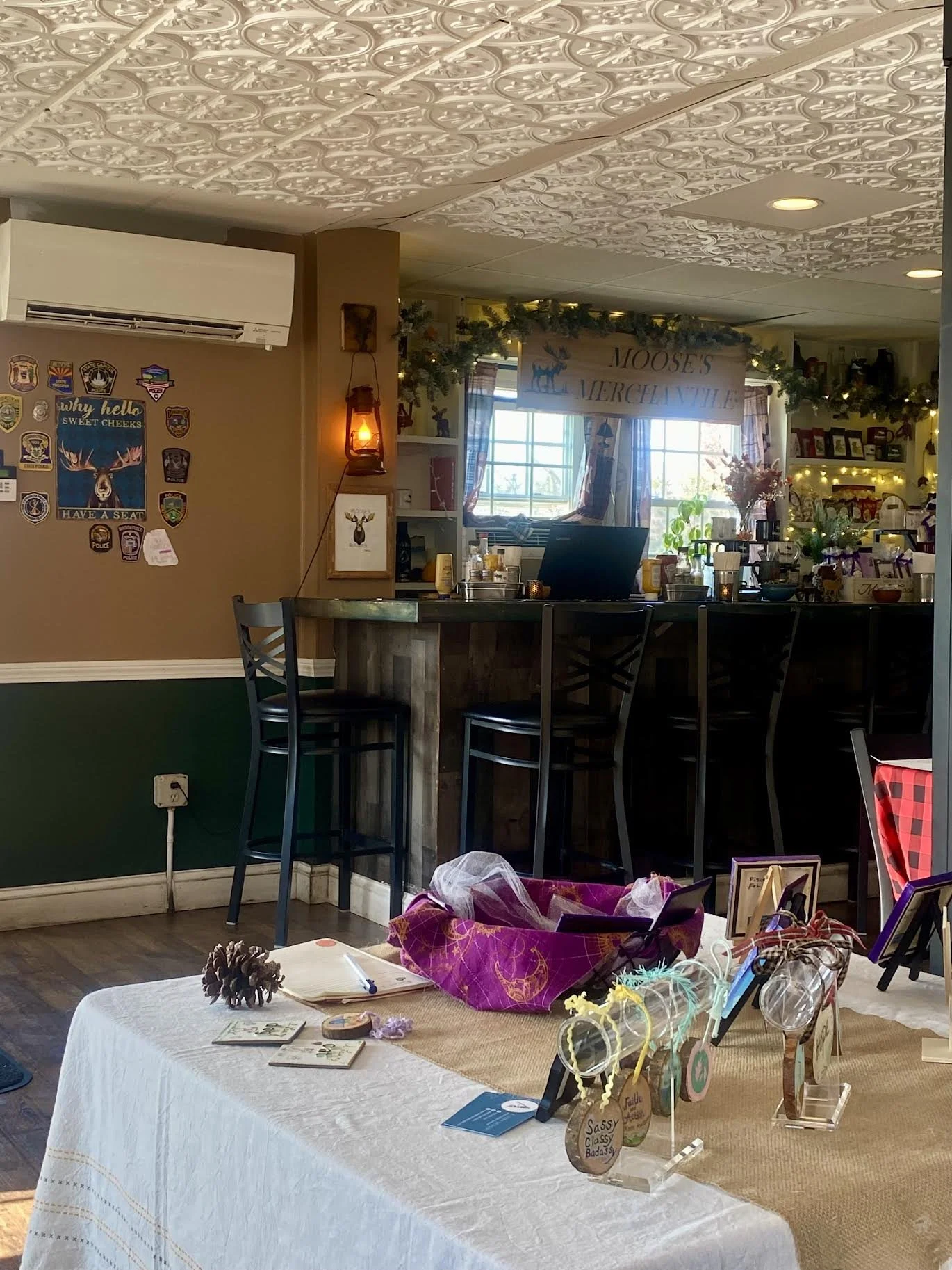 Interior of a cozy room with a table displaying small decorative items, framed photos, and gift bags. A bar area with four high chairs, a sign reading 'Moose's Mercantile,' and decorated windows are visible in the background.