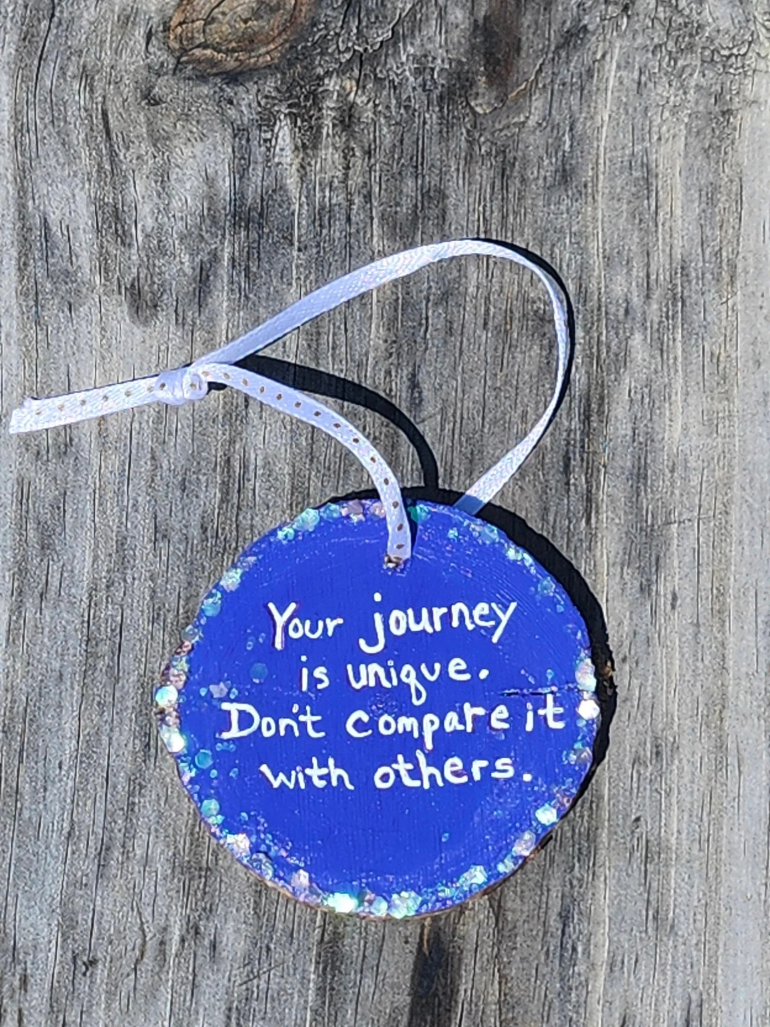 A circular blue ornament with white handwritten text that says, 'Your journey is unique. Don't compare it with others,' on a wooden surface with a white ribbon attached.