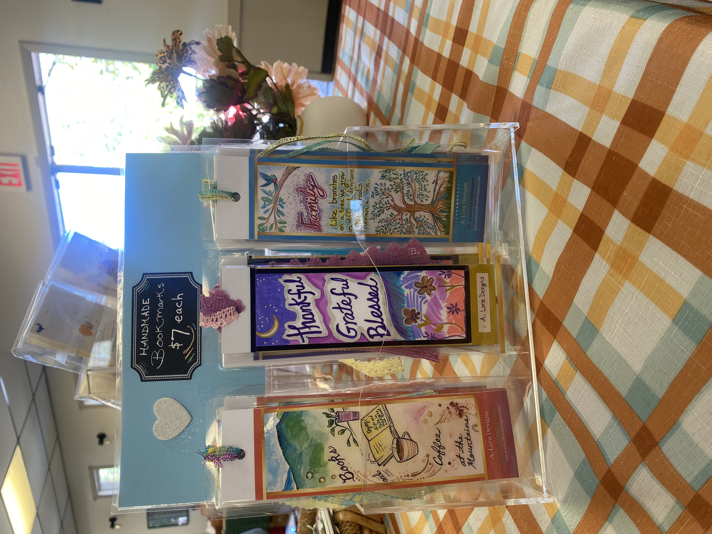 Clear acrylic display with handmade bookmarks inside, priced at $7 each. The bookmarks feature colorful watercolor designs with inspirational phrases such as 'Family', 'Like a breeze', and 'Thanks, grateful, blessed'. The display is decorated with he