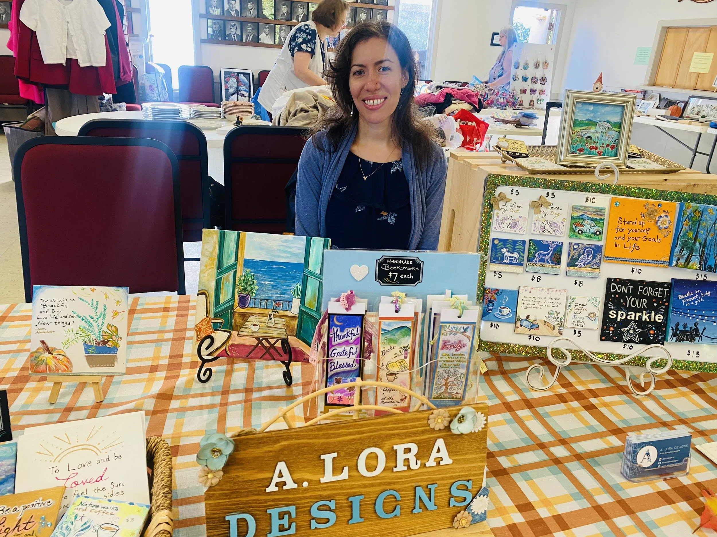 Woman sitting behind a table displaying various handmade art pieces and greeting cards at an indoor craft fair or market.