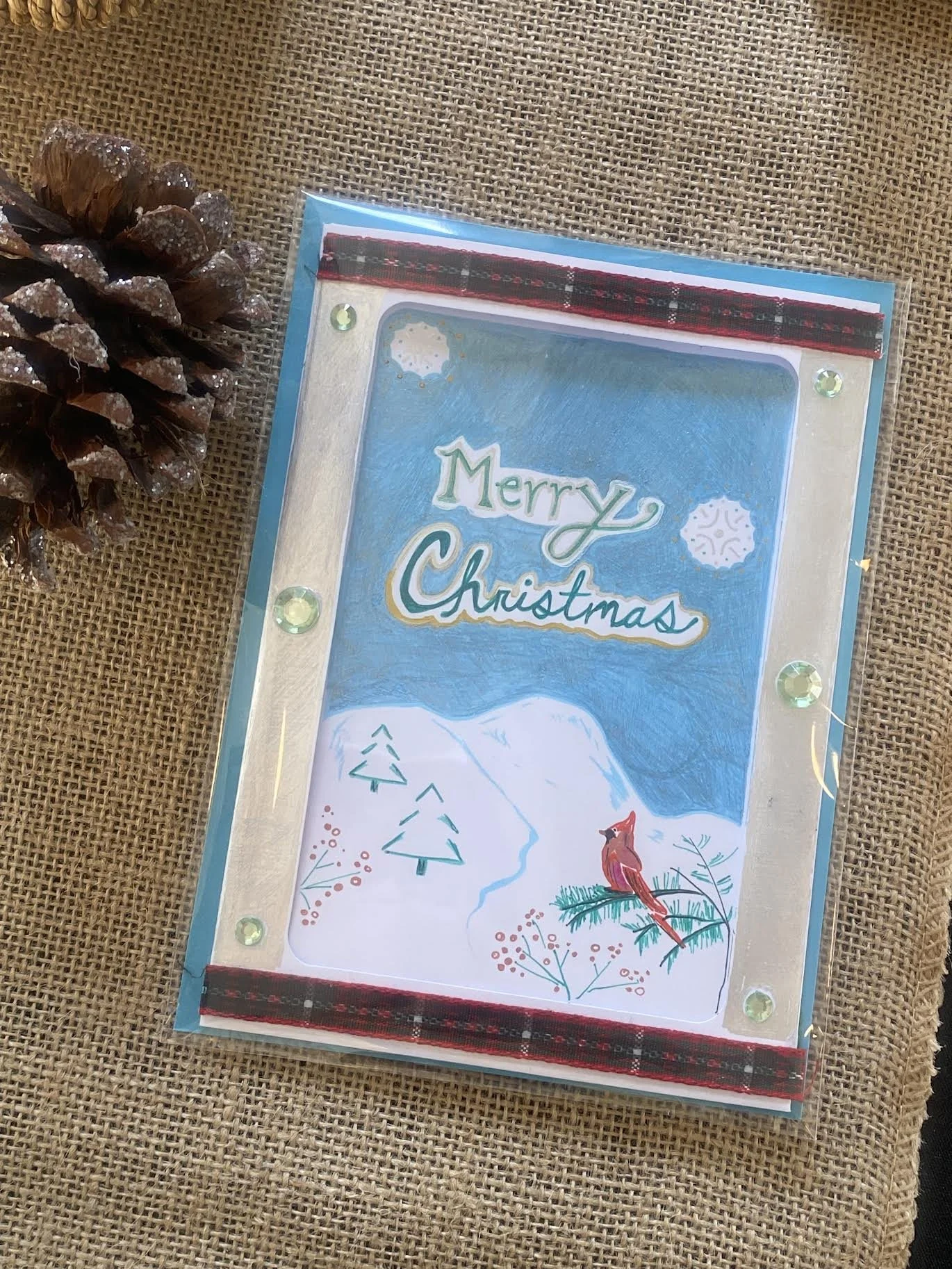 A Christmas card with a snowy mountain landscape, two pine trees, a cardinal bird, and snowflakes, with the words 'Merry Christmas' written on it. The card is decorated with green rhinestones and is inside a clear plastic sleeve, placed on a burlap s