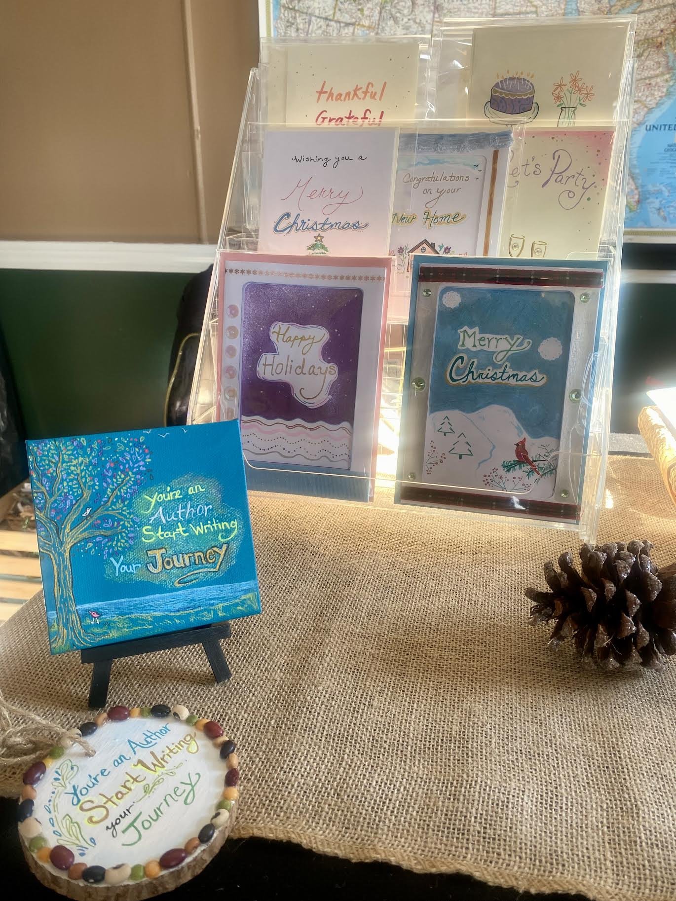 Display of holiday greeting cards, a small painting with motivational quote, and a pine cone on a burlap-covered table.