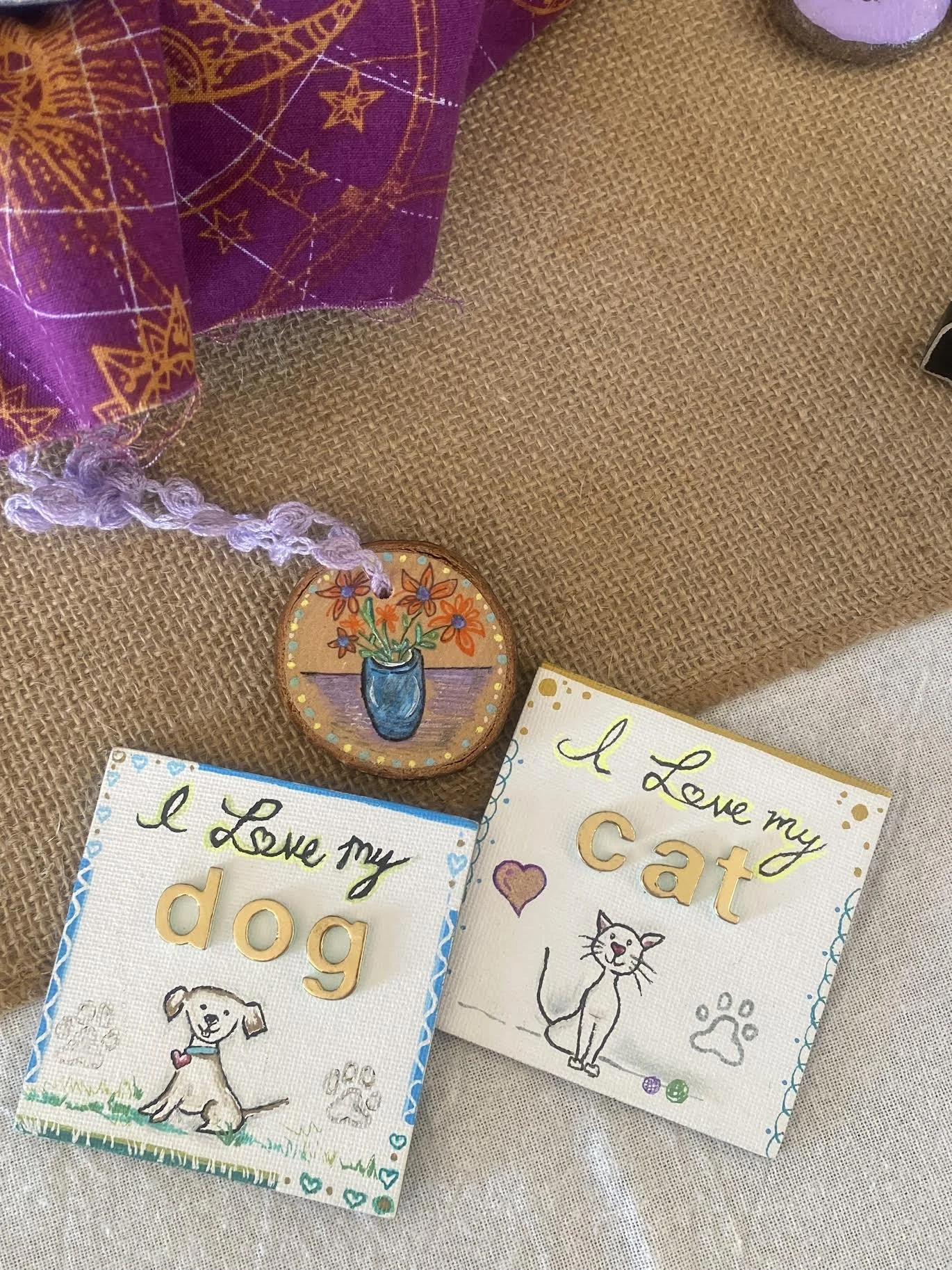 Handmade signs expressing love for a dog and a cat, with illustrations of each pet, and a painted wooden ornament of a vase with flowers, all placed on a brown and white fabric surface.