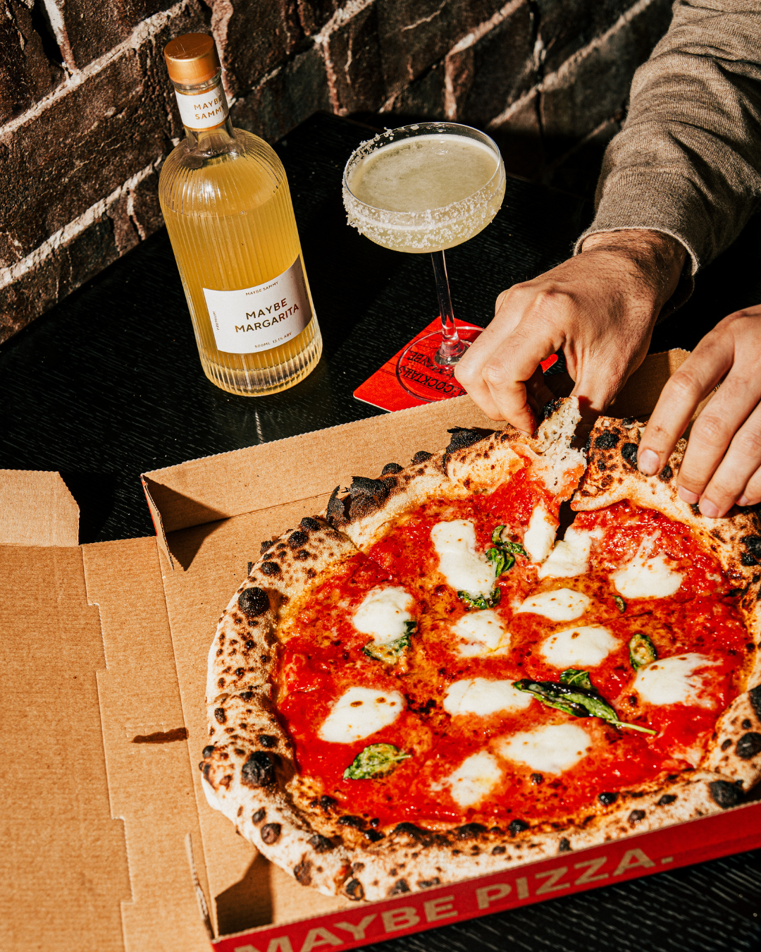 Close-up of a hand picking up a slice of Margherita pizza from an open pizza box on a black table, with an additional bottle of Maybe Margarita cocktail and a glass of cocktail in the background against a brick wall.