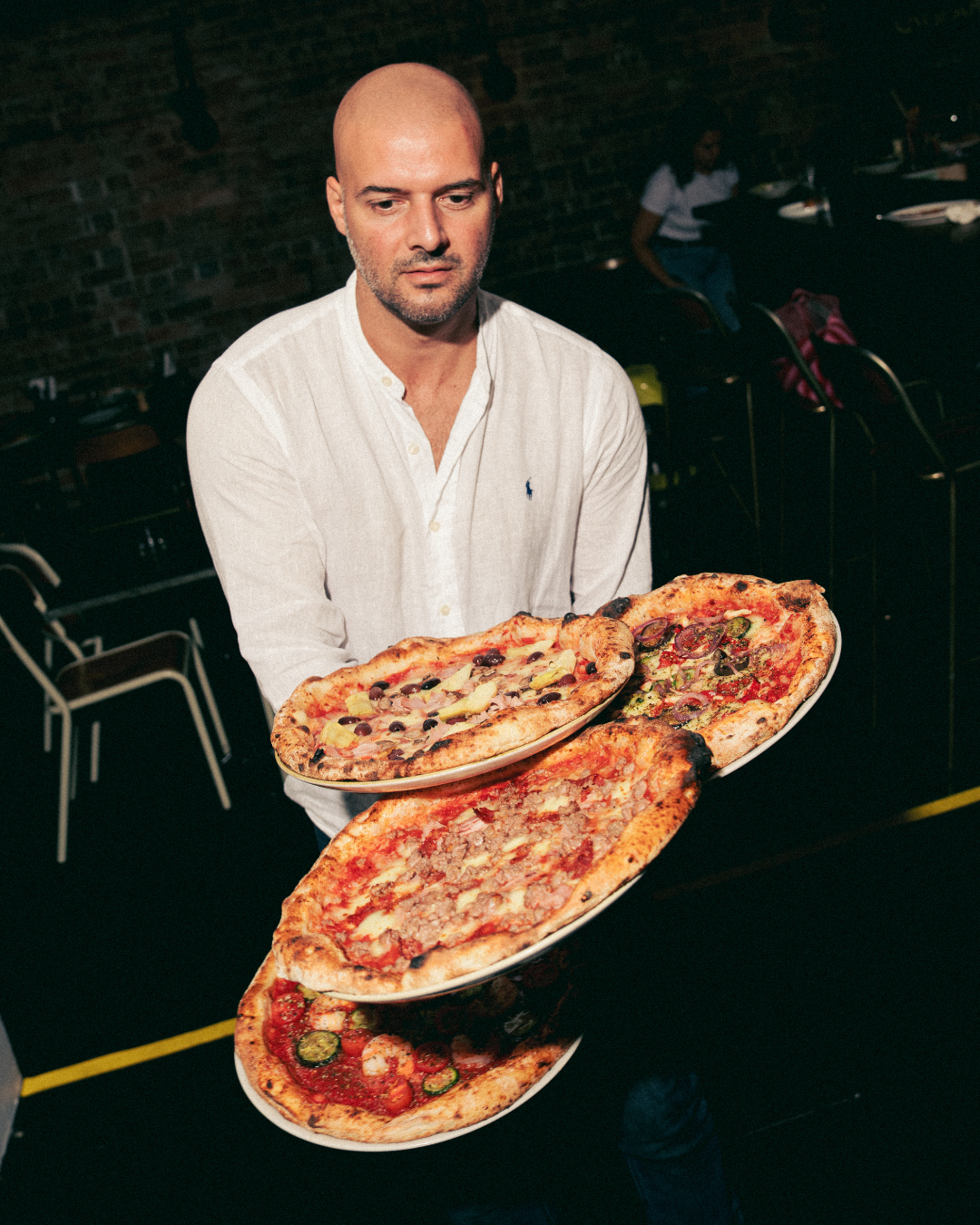 A man in a white shirt holding four large plates with different types of pizza inside a restaurant with a dark brick wall.