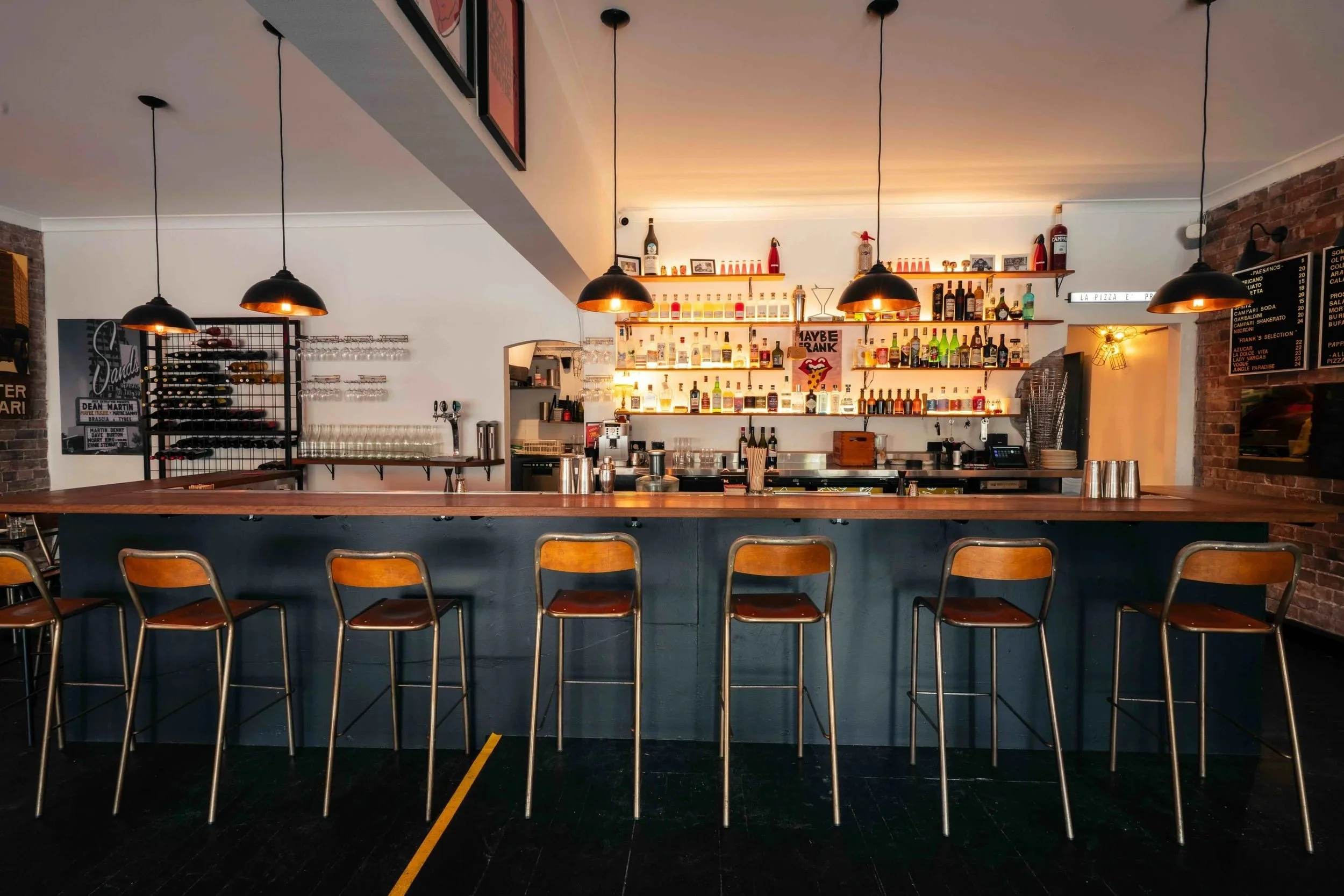 Interior of a bar with a long counter, wooden barstools, dim pendant lighting, shelves with liquor bottles, and a chalkboard menu on the wall.