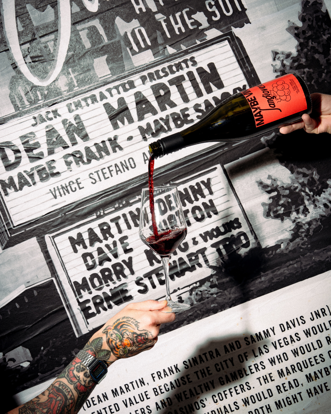 Person pouring red wine into a wine glass, with tattoos on their arm and wearing a smartwatch, against a background of black and white posters for music albums and concerts.