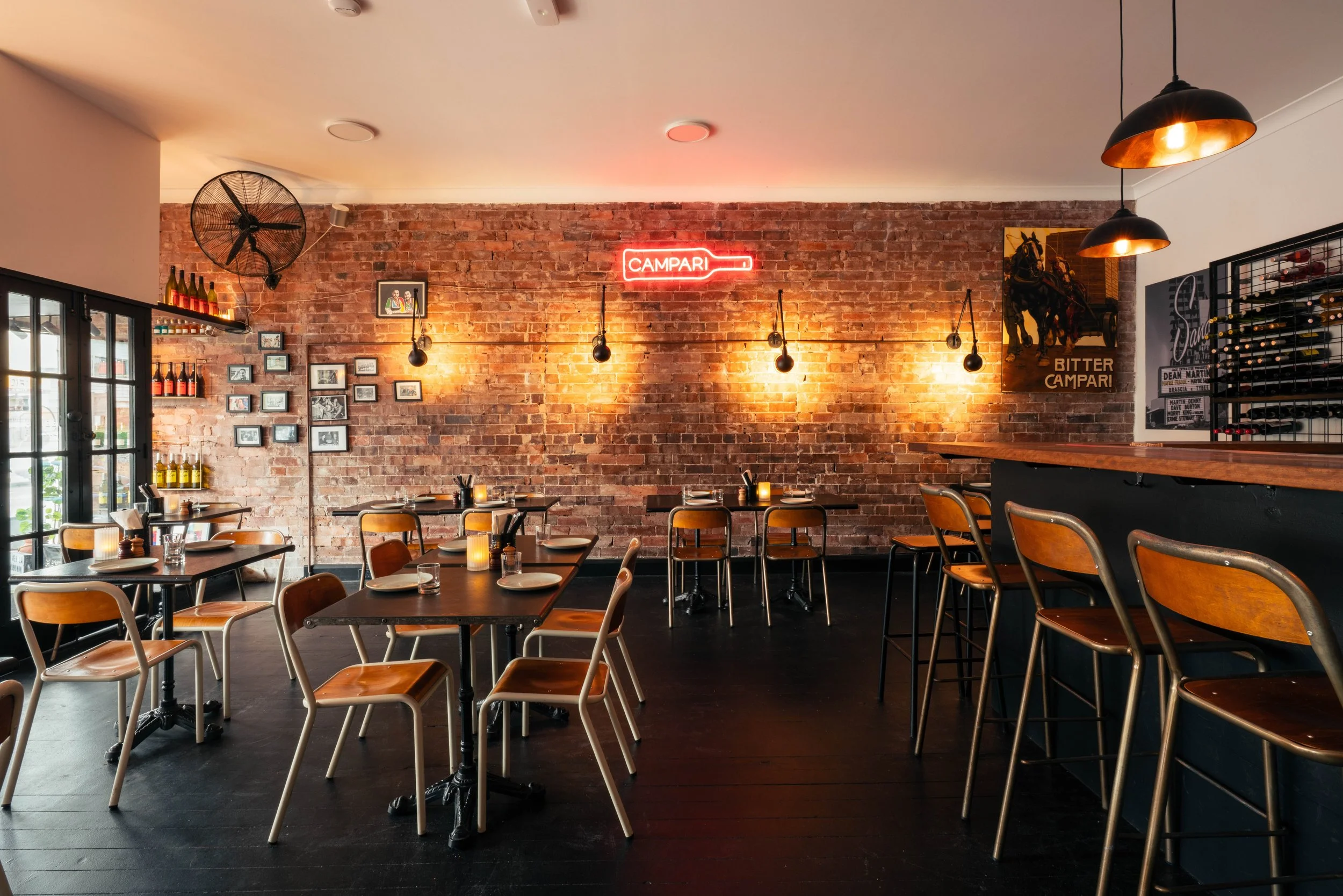 Interior of a restaurant or cafe with wooden tables and chairs, exposed brick wall, hanging pendant lights, and a neon sign that says "CAMPARI". Decor includes photos on the wall, wine bottles on a shelf, and artwork.