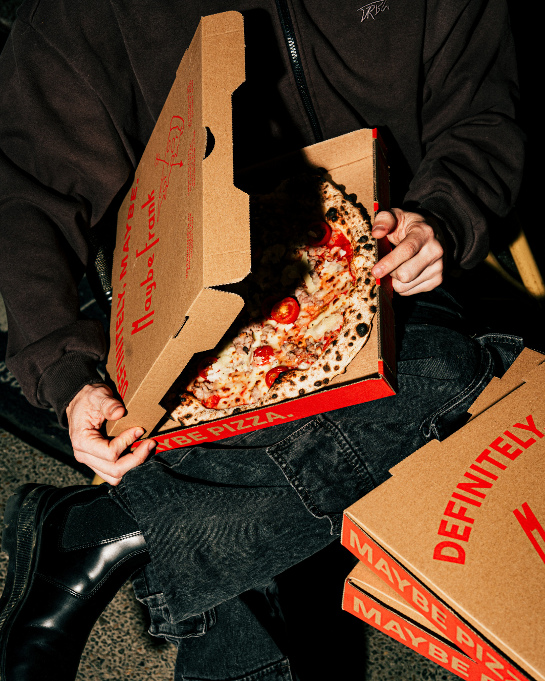 Person holding an open pizza box with a pepperoni pizza inside, sitting on a chair with several closed pizza boxes stacked nearby.