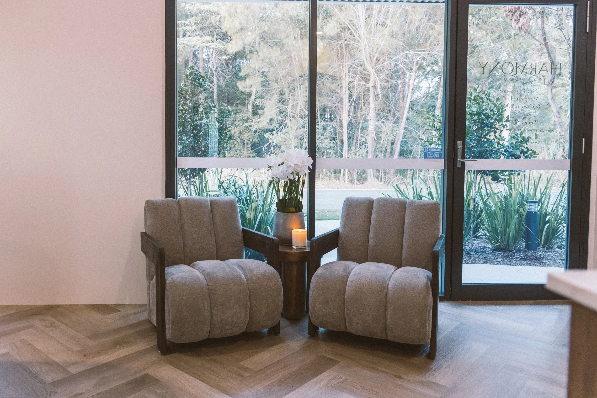 Two gray upholstered chairs face each other with a small wooden side table between them. On the table is a white orchid plant in a gray pot and a lit candle. Behind the chairs is a large glass door with a view of green plants and trees outside.