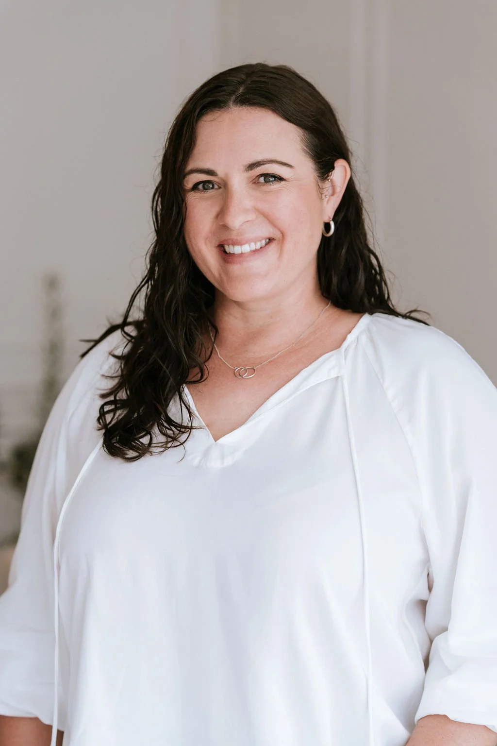 A woman with dark brown, curly hair wearing a beige and red striped blouse standing against a plain light-colored wall.