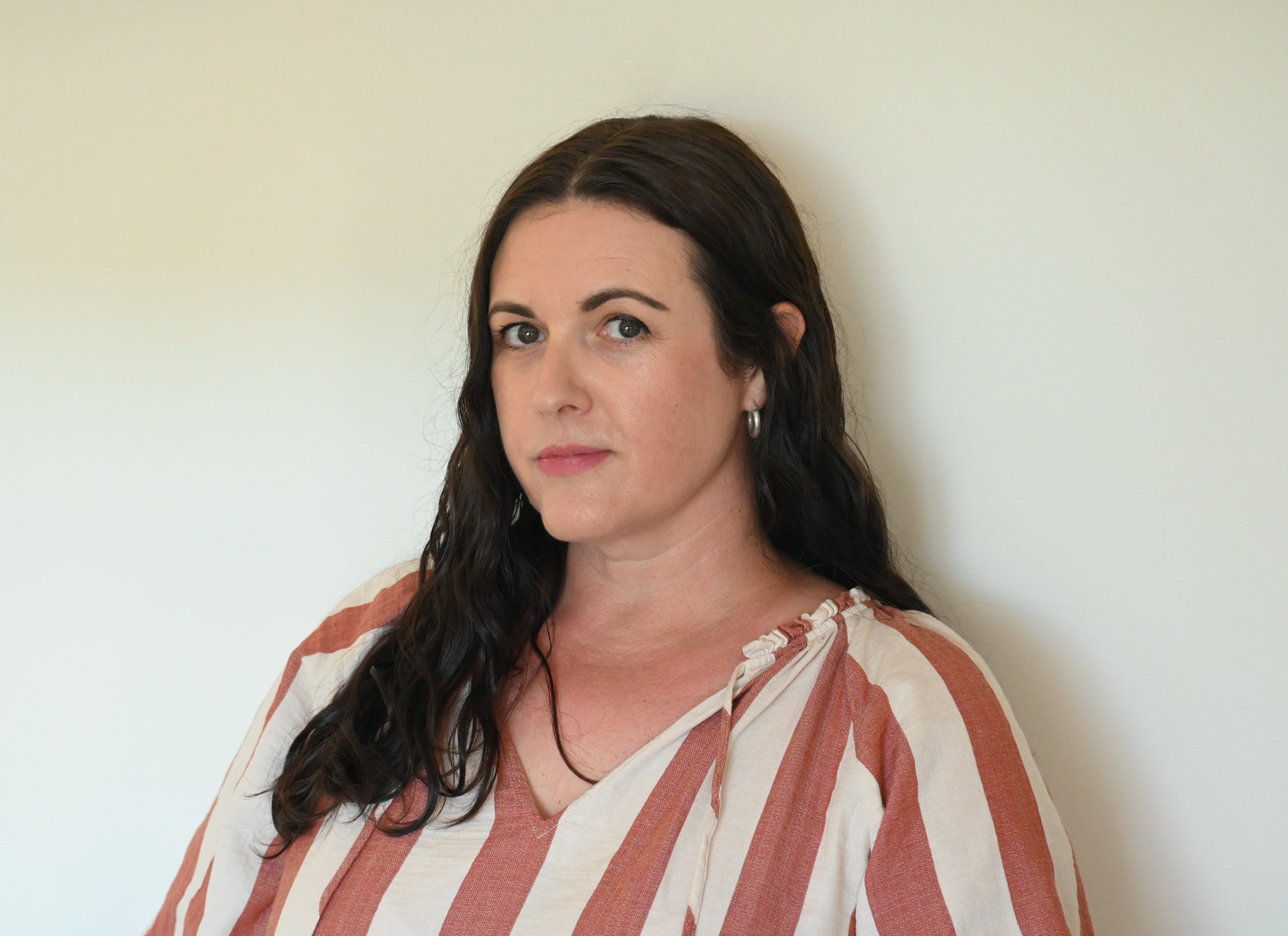 A woman with long dark wavy hair and light skin, wearing a cream and rust striped blouse, standing against a plain off-white wall.