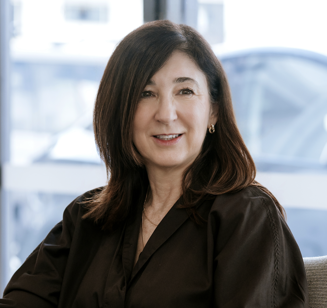 A middle-aged woman with dark brown hair and hoop earrings, smiling, wearing a beige button-up shirt, sitting in a bright room with white walls and a potted plant in the background.