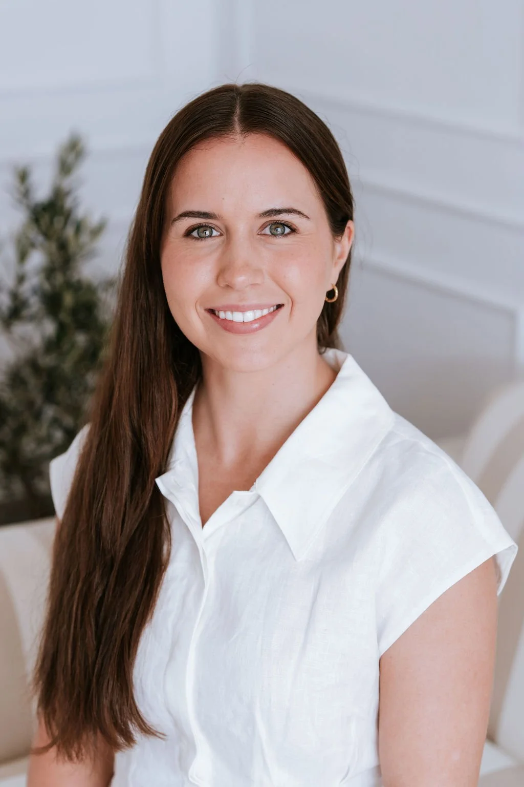 A woman with long brown hair, green eyes, and a white shirt smiling at the camera, in a bright, white room with a blurred plant in the background.