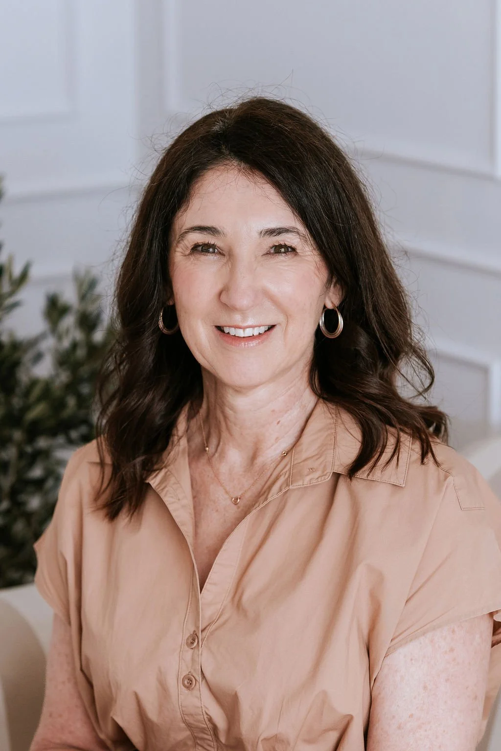 A middle-aged woman with dark brown hair and hoop earrings, smiling, wearing a beige button-up shirt, sitting in a bright room with white walls and a potted plant in the background.