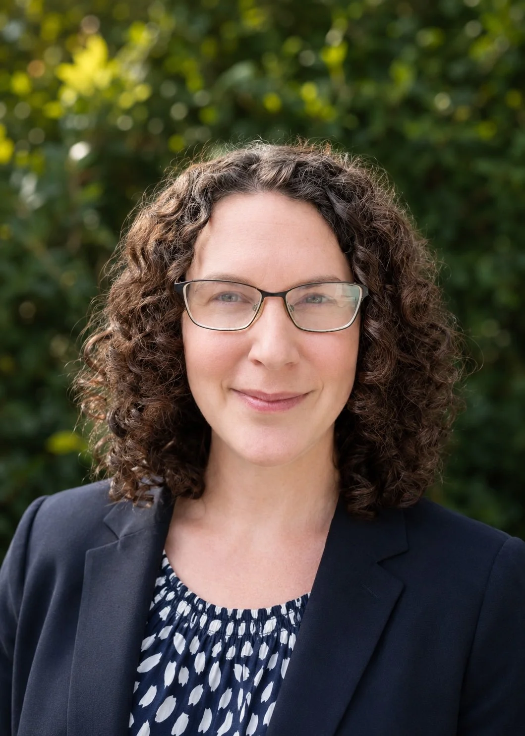 A woman with curly brown hair and glasses smiling outdoors in front of green shrubbery.