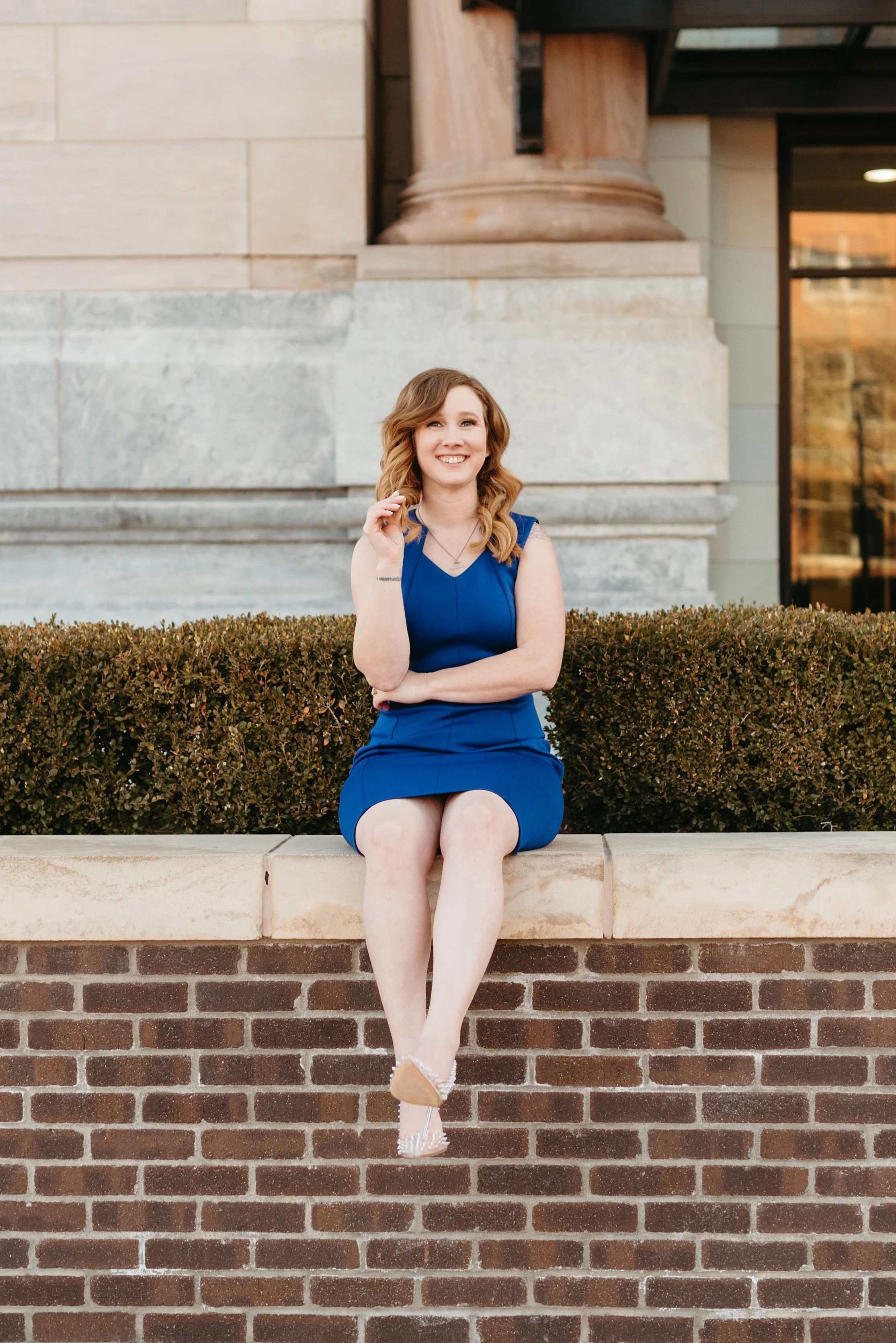 Woman in blue dress sitting on a brick wall, smiling, with decorative pillars in the background.