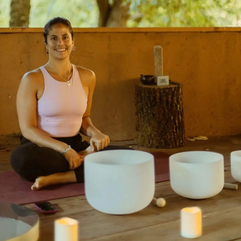 Person sitting cross-legged on a cushion with a singing bowl and mallet nearby, practicing meditation outdoors.