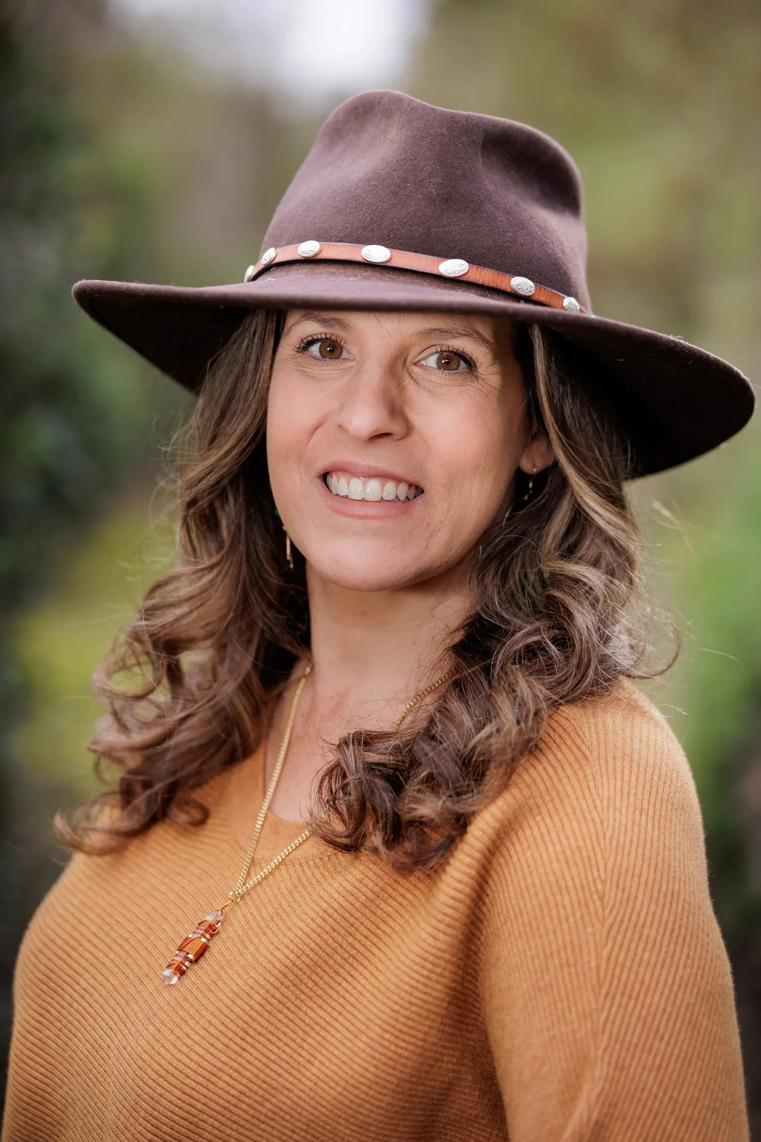A woman with brown curly hair wearing a wide-brimmed brown hat with decorative silver conchos, a mustard-colored sweater, and a gold necklace with a pendant, outdoors with blurred green foliage in the background.