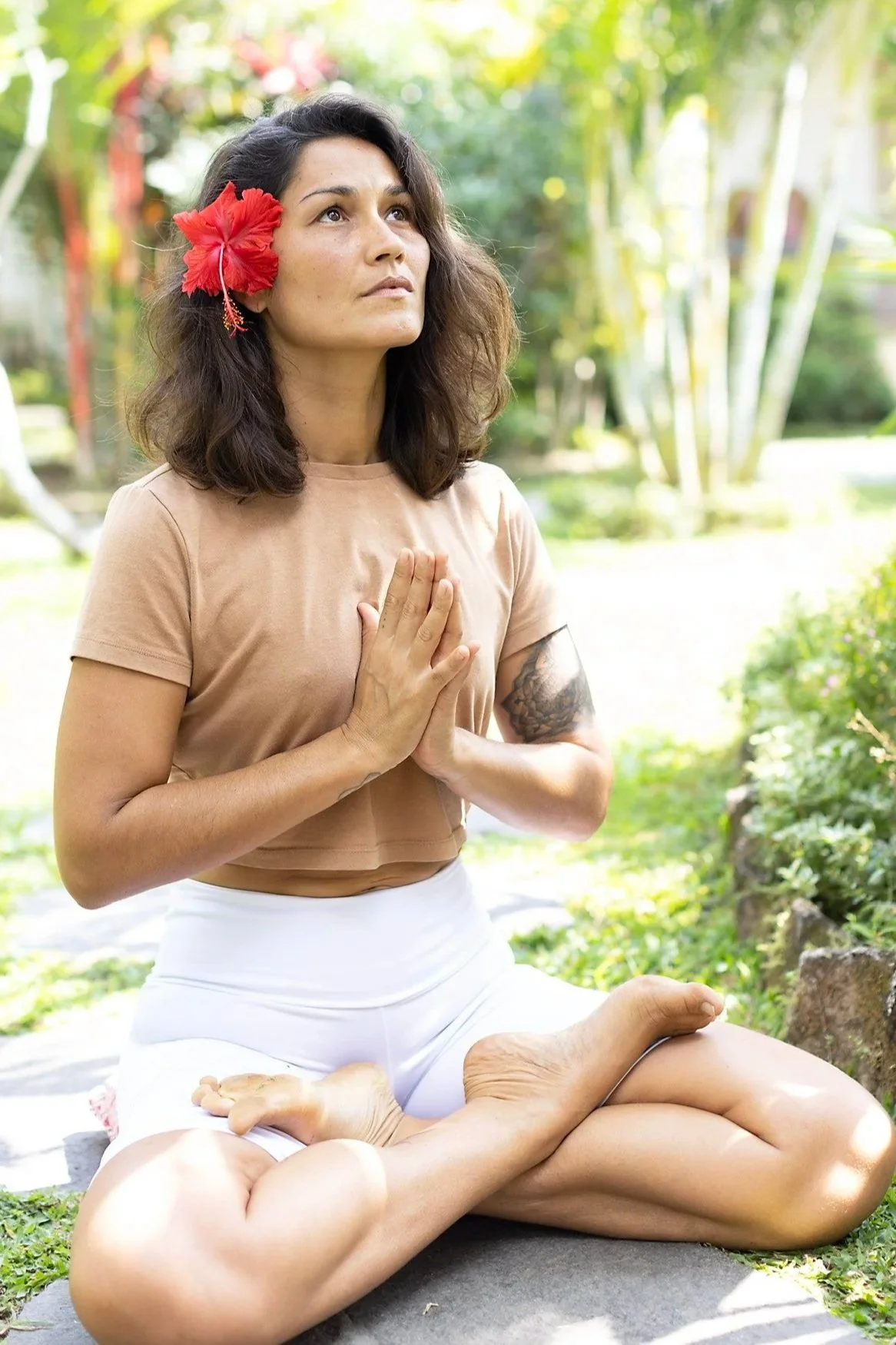 Person smiling in a white tank top with a colorful floral tattoo on their arm, standing outdoors on a forest path.