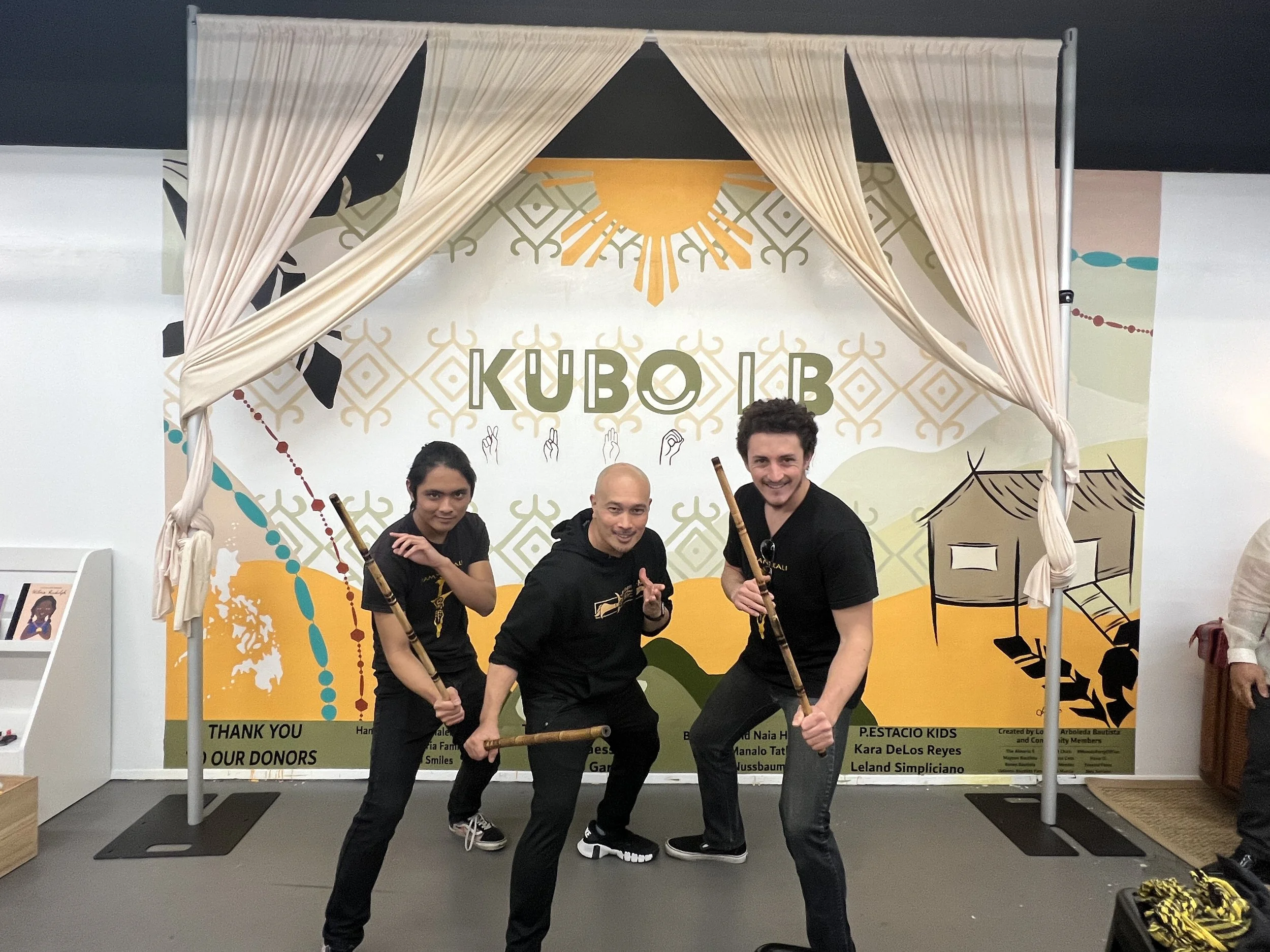 Three men posing with props in front of a colorful backdrop at an event. The men are smiling and holding sticks, dressed in black. The backdrop displays the word 'KUBO' with decorative patterns and a thank you note at the bottom.