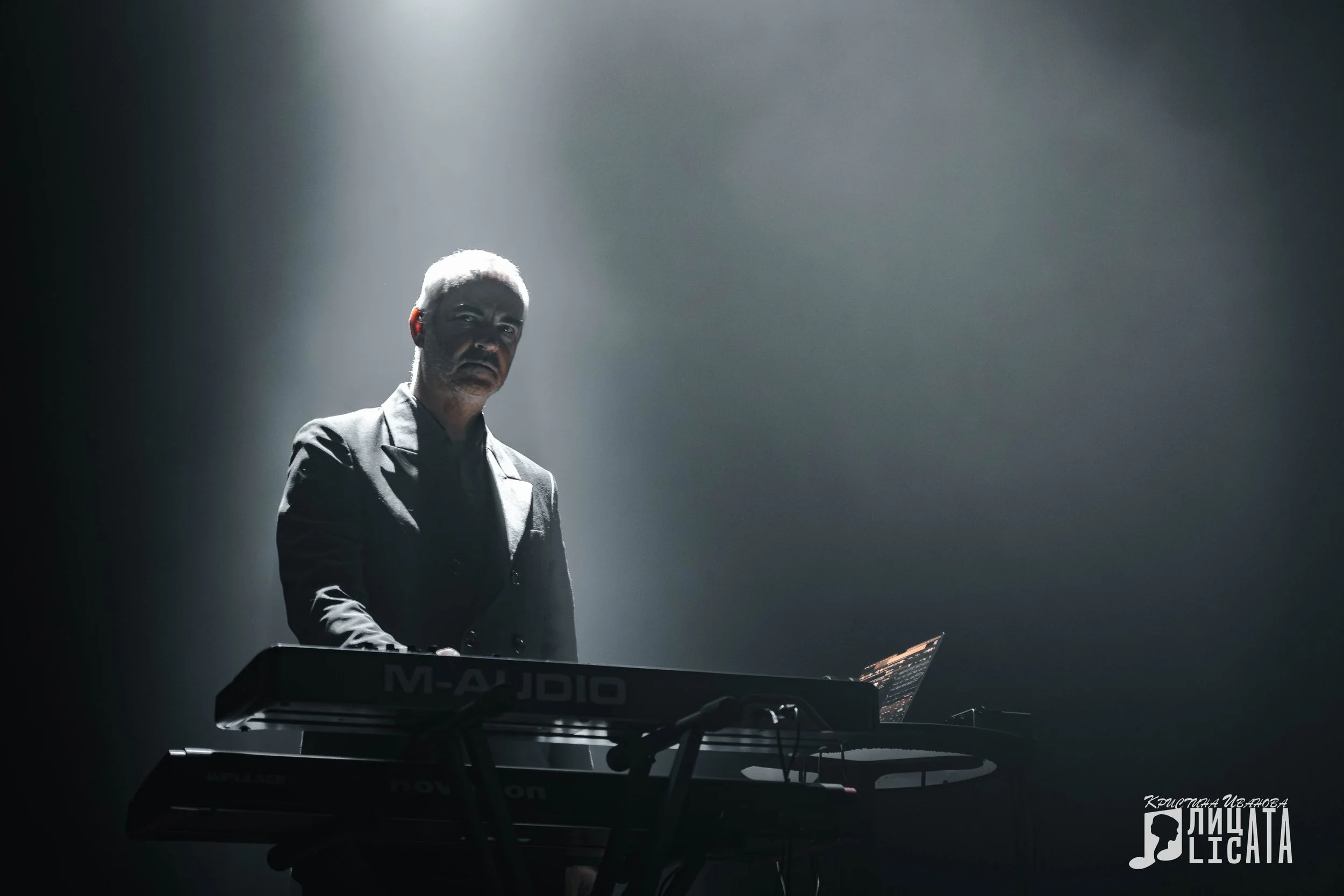 A man with gray hair wearing a dark suit, standing in a spotlight, playing a keyboard on a dark stage.