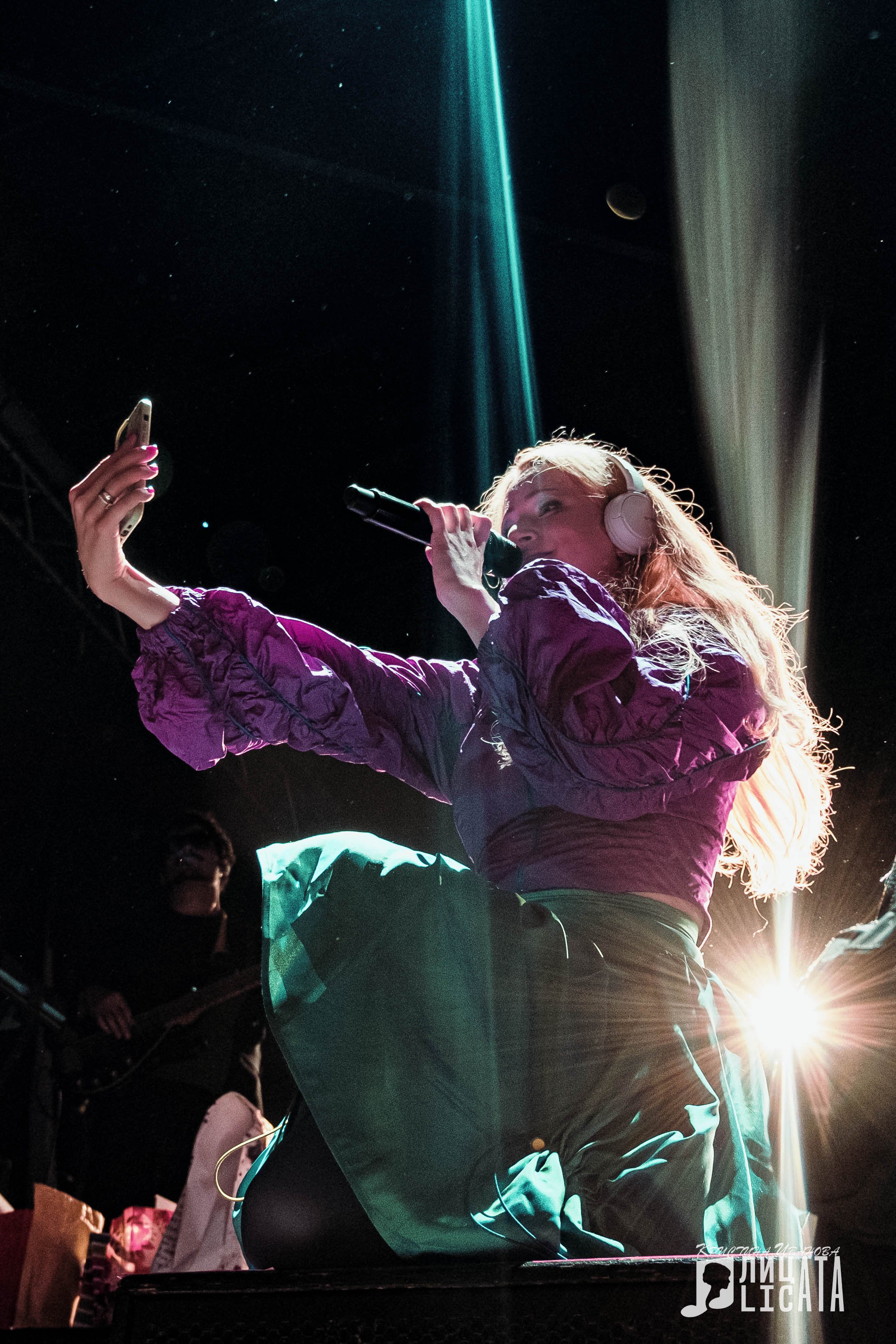 A female singer with pink headphones singing into a microphone on stage, taking a selfie with her smartphone, with stage lights and a band member in the background.
