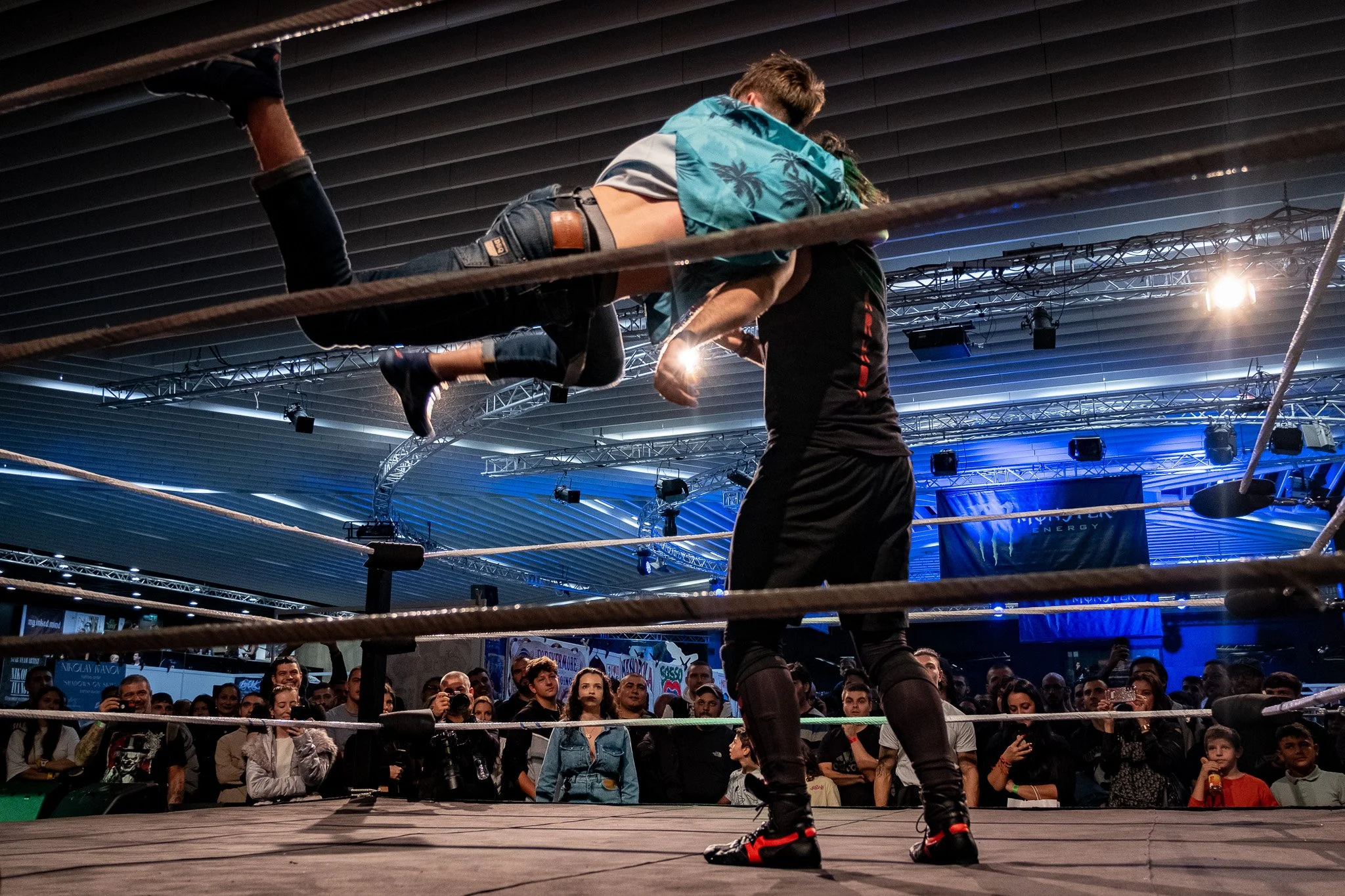 A professional wrestling match taking place indoors with a crowd watching as a wrestler is elevated over the ring ropes by a referee or trainer.