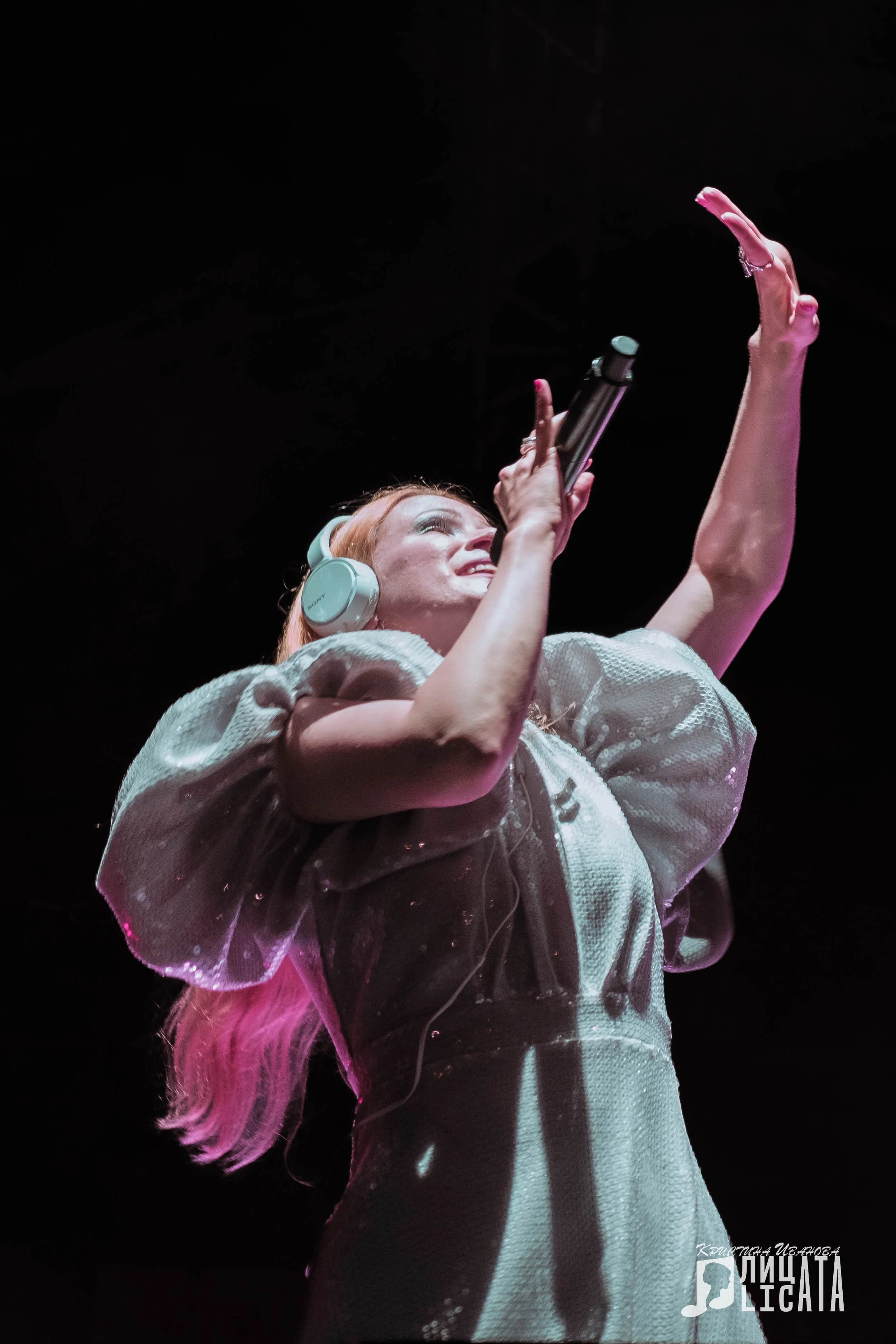 A woman singing on stage with headphones, holding a microphone, and raising her hand.