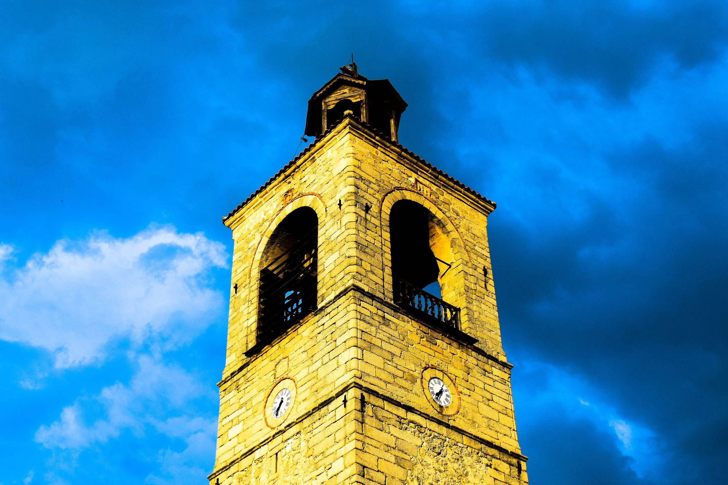 A historic yellow stone bell tower with a clock, set against a stormy blue sky.