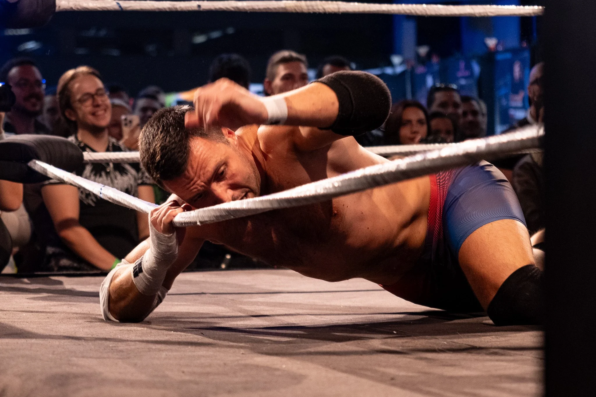 A male wrestler is lying on the mat next to the ropes inside a wrestling ring, with a pained expression on his face, surrounded by an audience watching the match.