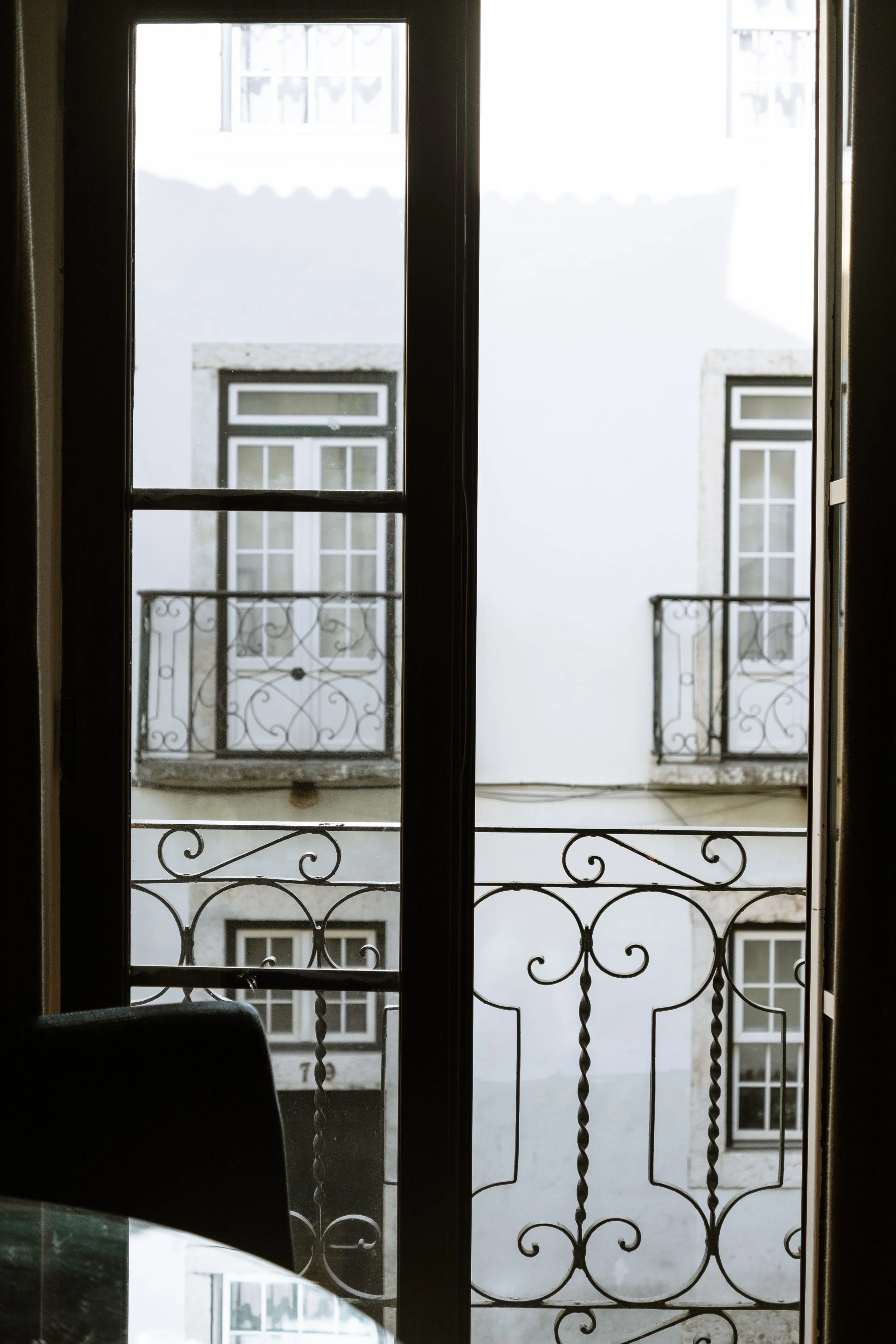 Image shows a view through a glass door with an iron balcony railing, overlooking a white building with multiple windows.