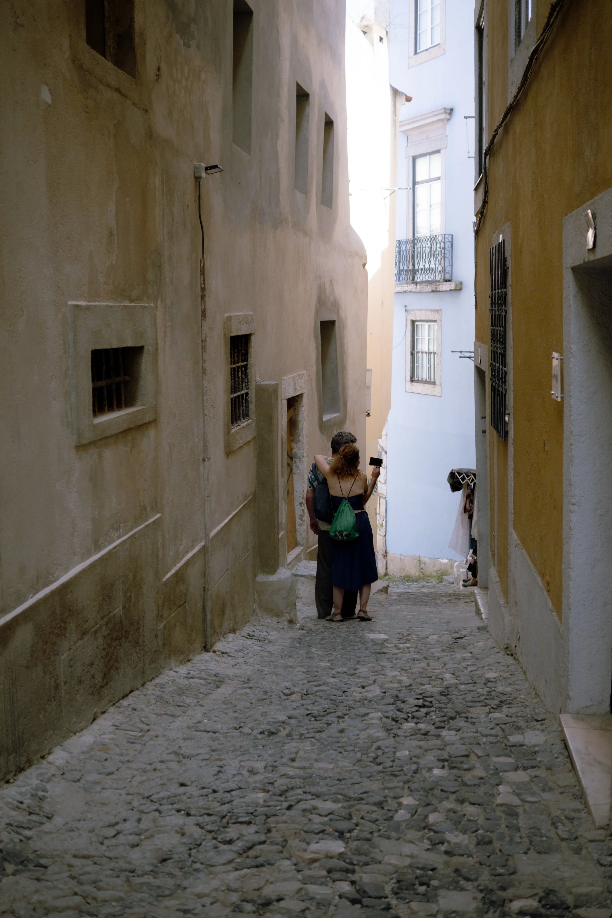Two people standing close together and looking at a smartphone in a narrow cobblestone alleyway with old buildings on either side.