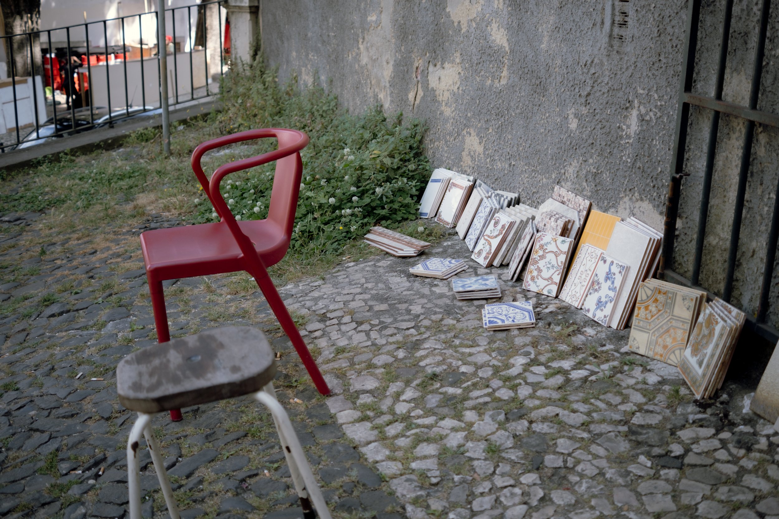 Outdoor scene with a red chair, a stool in front, and a display of patterned ceramic tiles leaning against a wall.