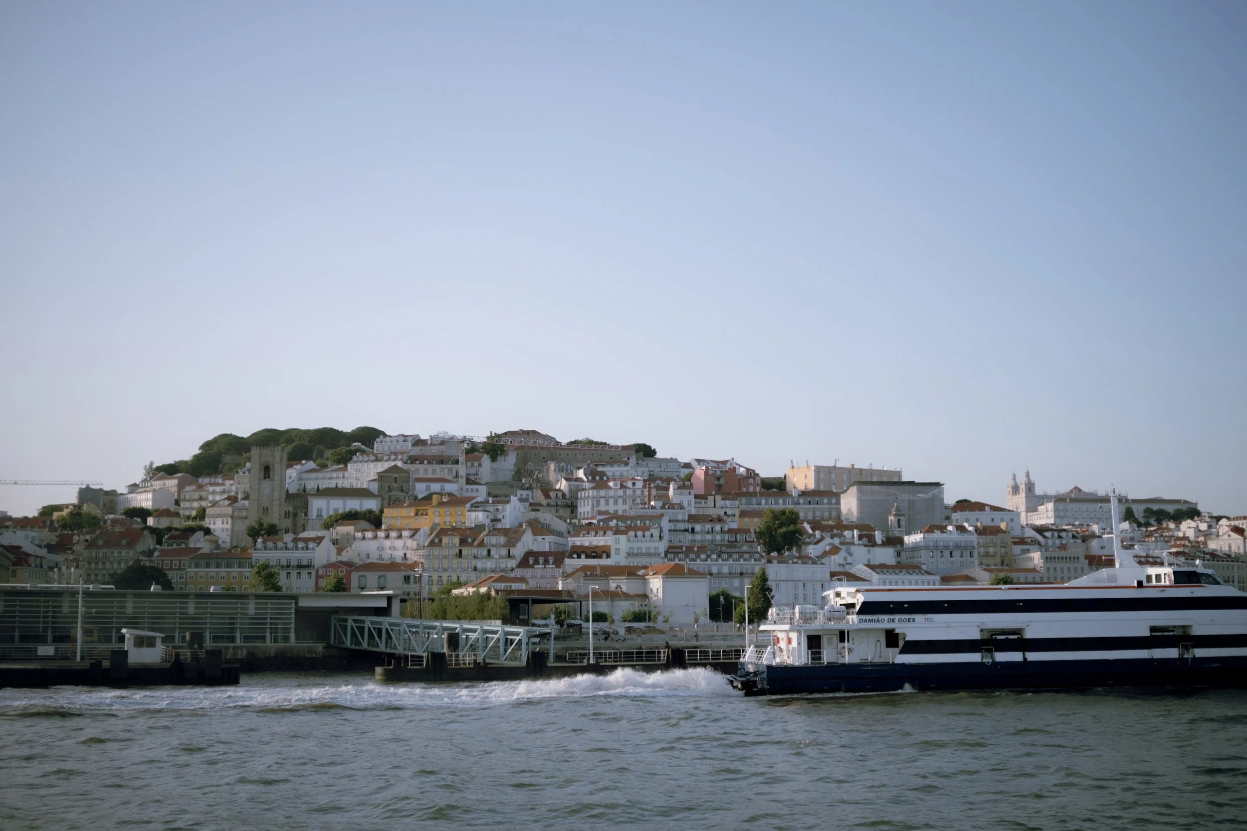 A view of a hillside cityscape with white buildings, a church tower, and a large ferry on a river in the foreground.