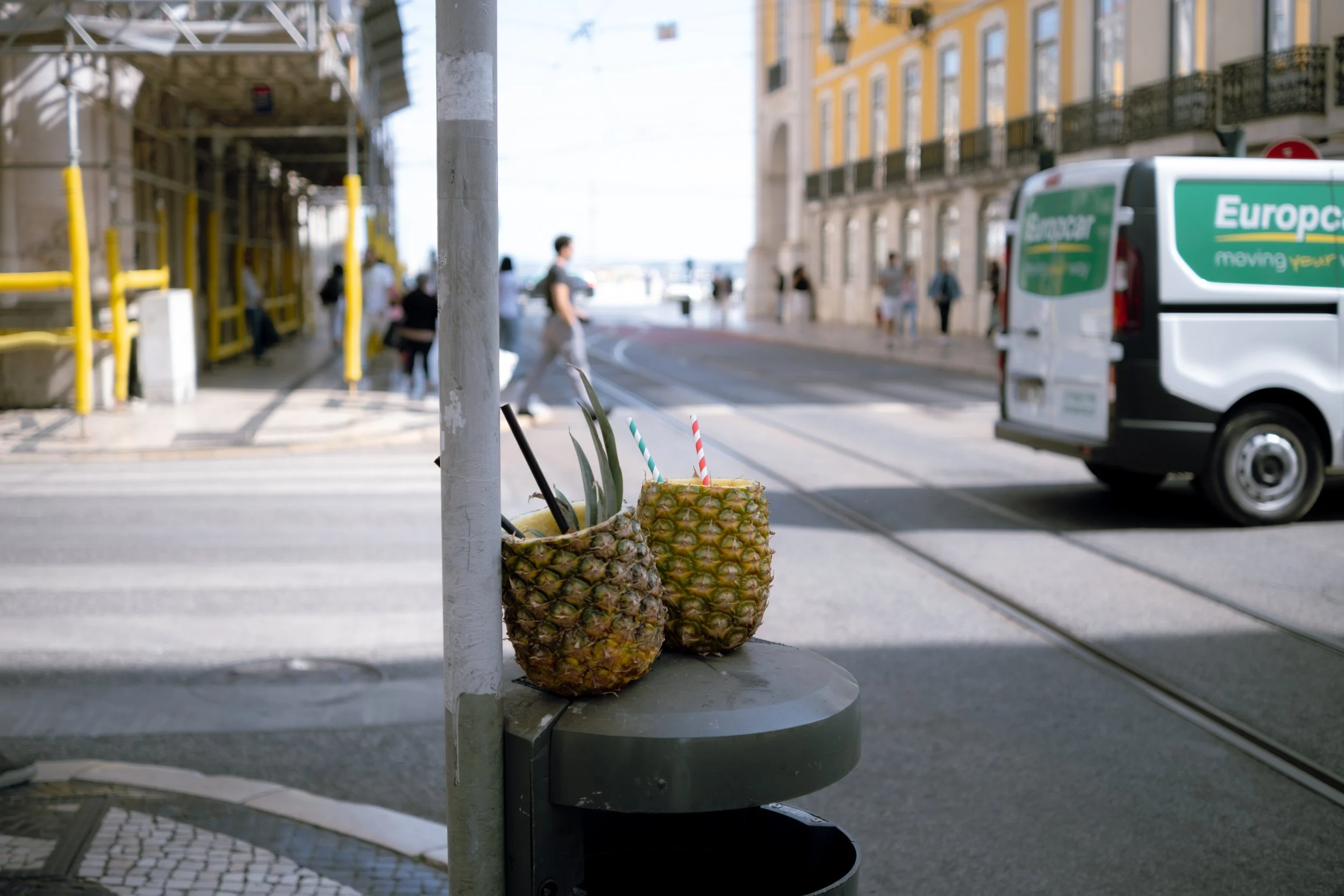 Two pineapples with cutouts and straws used as drinks sit on a small table on a city street, with tram tracks, a white van, and blurred pedestrians in the background.