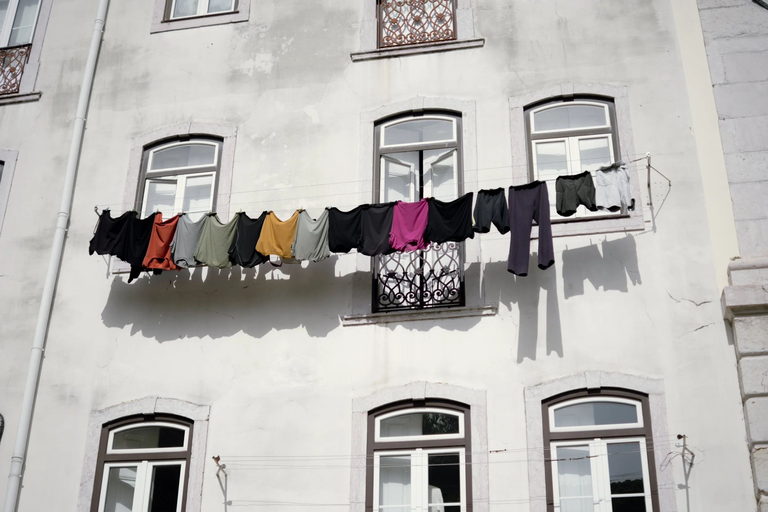 Clothes hanging on a clothesline in front of a building with white walls and arched windows, casting shadows.